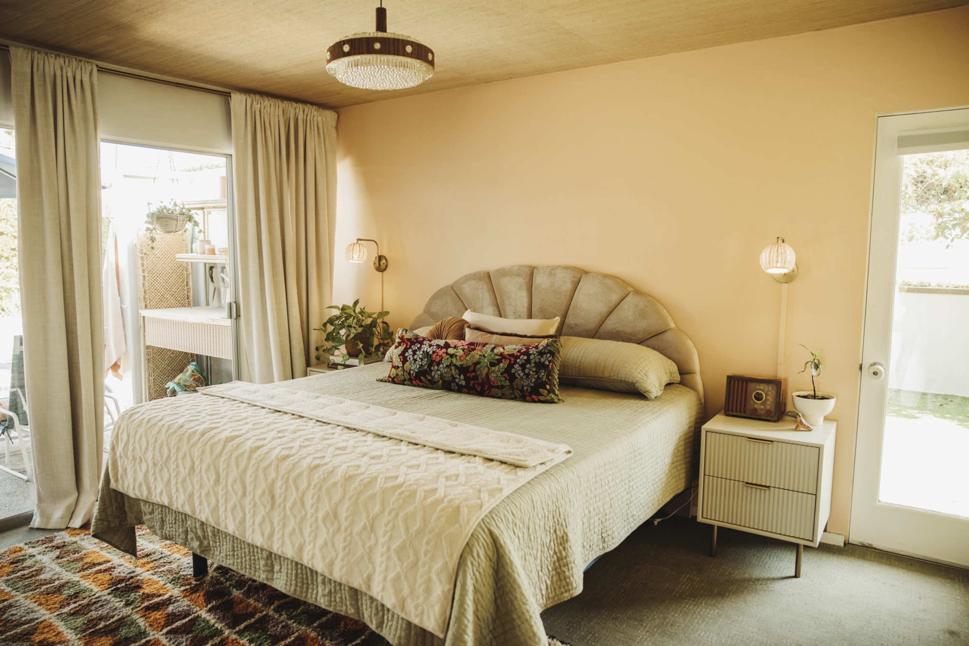 The image shows a neatly arranged bedroom with a large bed, a patterned rug, and a sliding glass door leading to an outdoor space.
