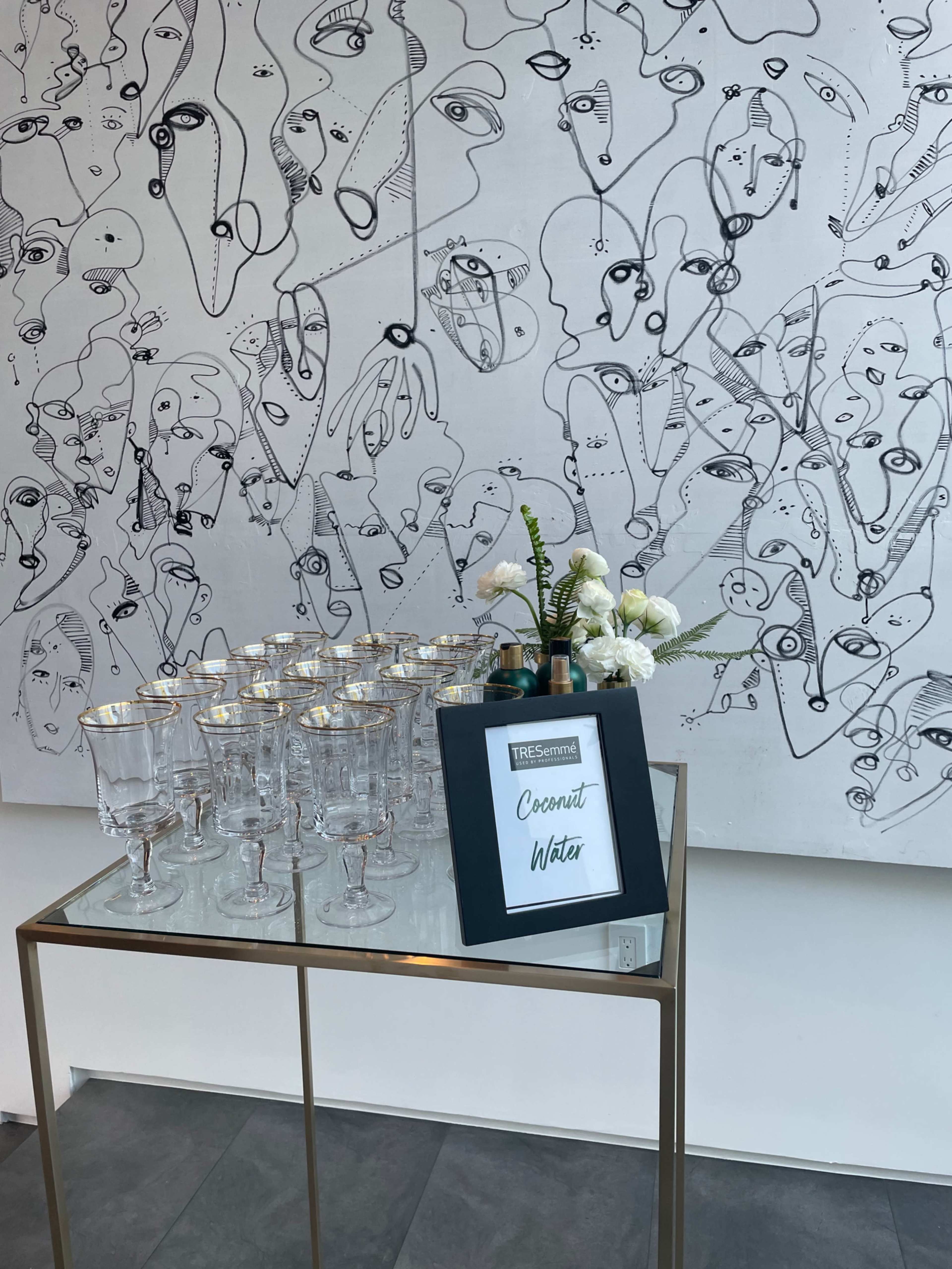A table displays crystal glasses alongside a sign for coconut water, set against a large abstract line drawing.