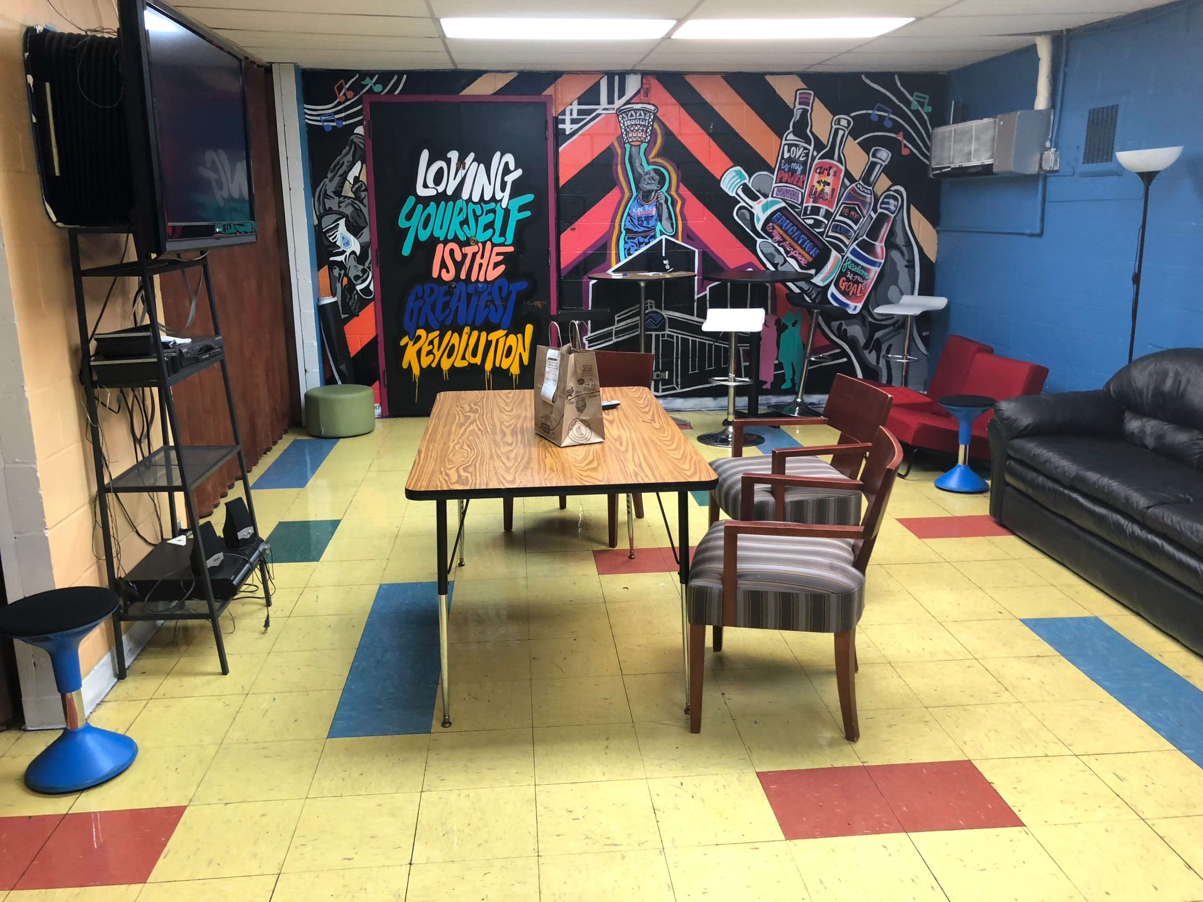 The room features a table with chairs, a leather couch, and a colorful mural on the wall, along with a television and other furniture.