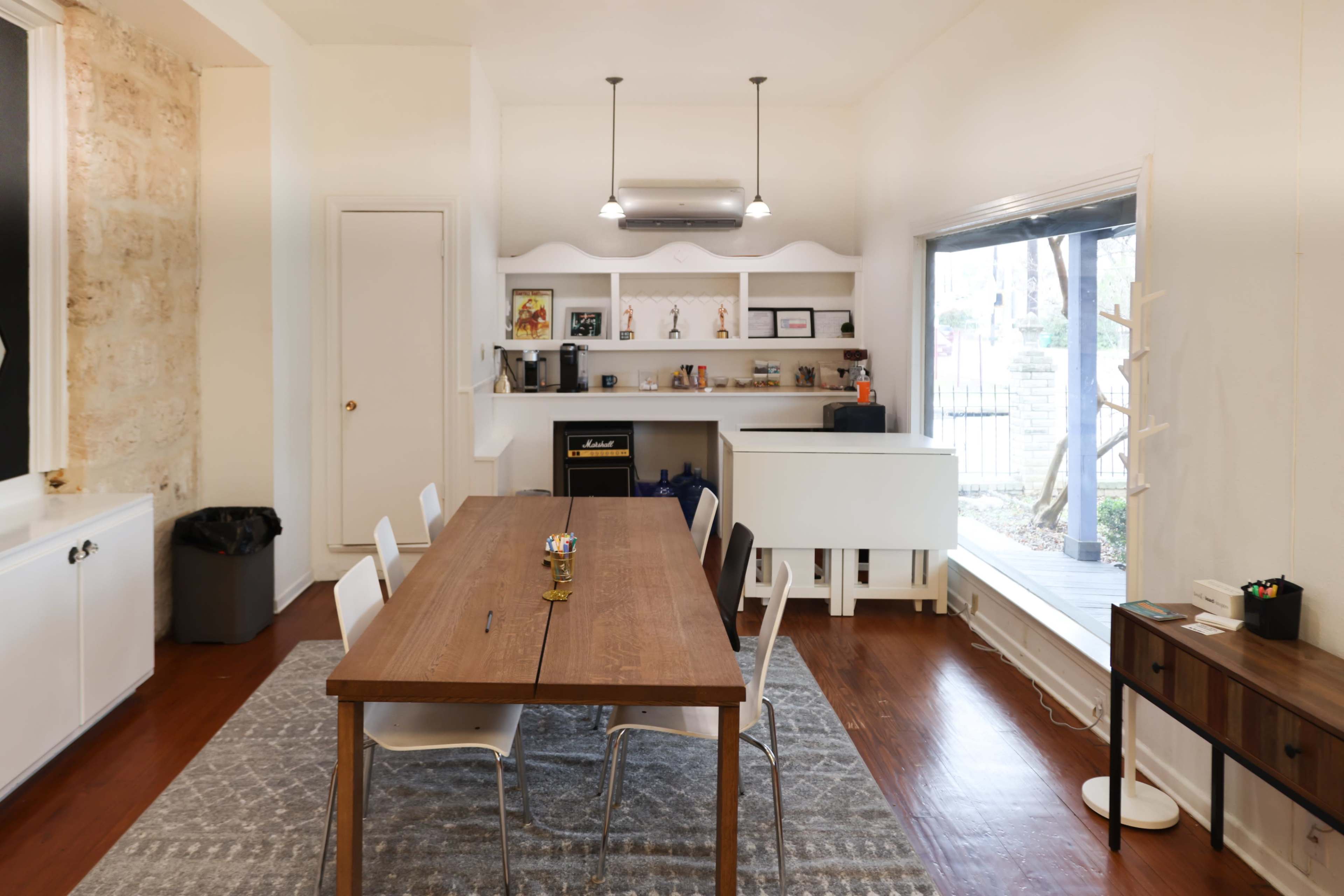 A wooden dining table with white chairs is positioned in a brightly lit room featuring a small kitchen area and large windows.