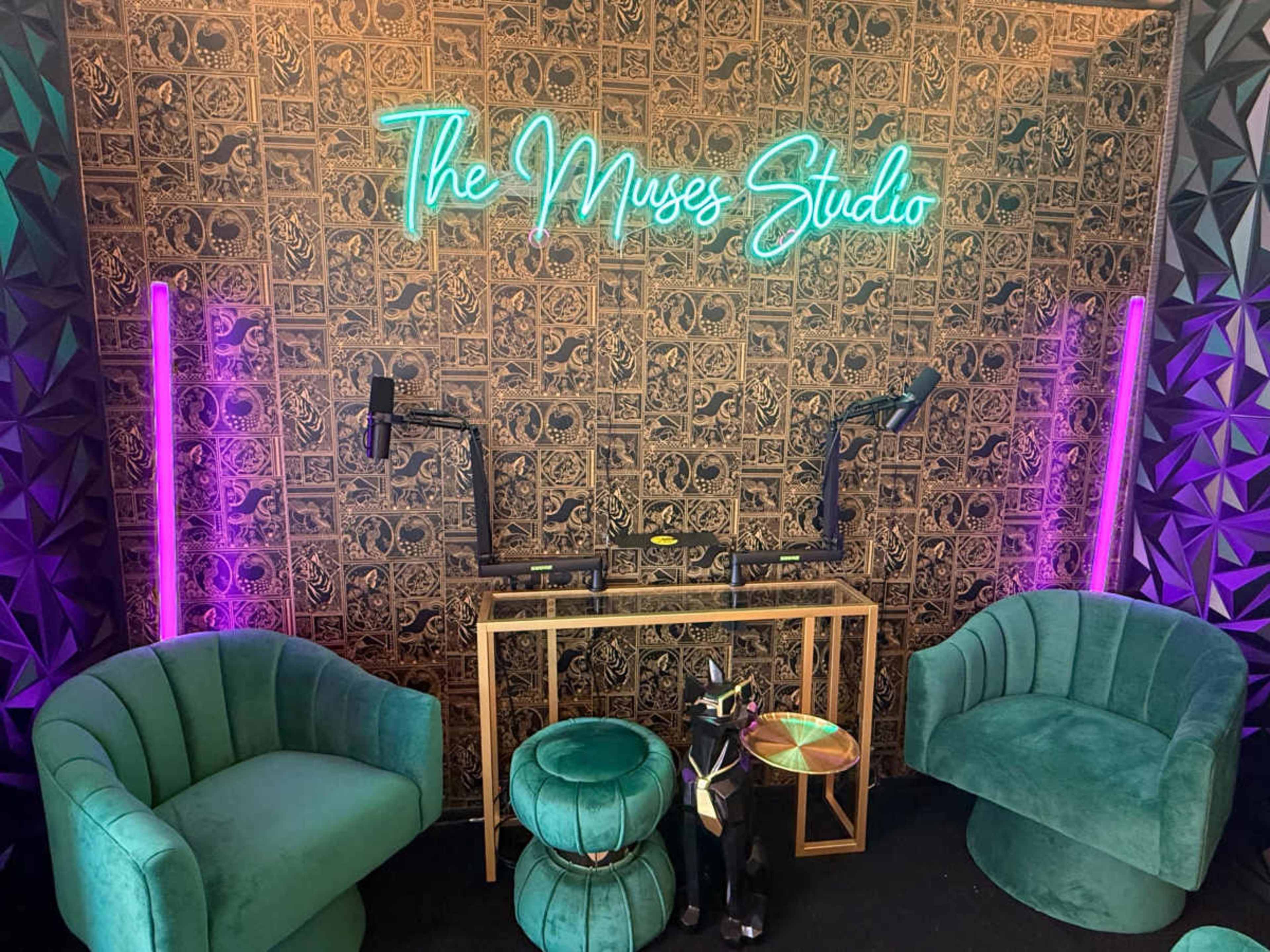The image shows a modern podcasting studio featuring two teal chairs, a small table, a neon sign that reads "The Muses Studio," and two microphone setups against a patterned wall.