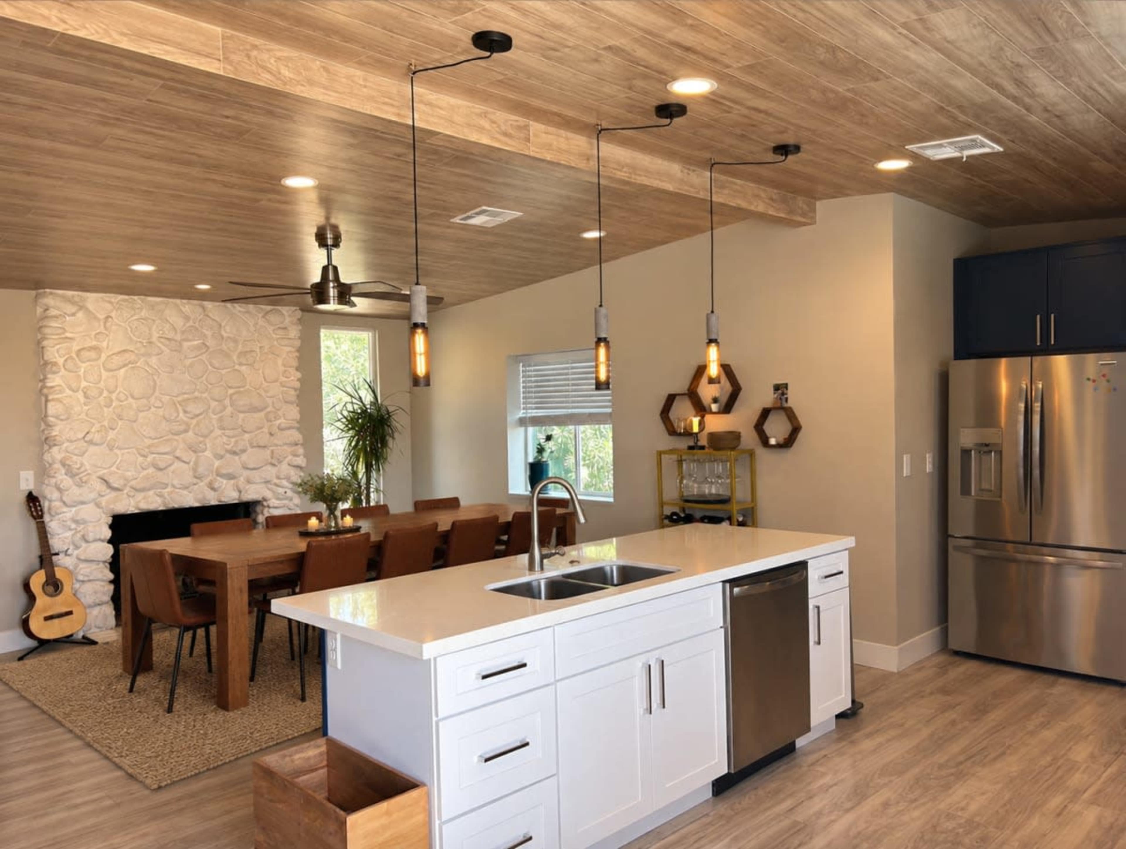 The image shows a modern kitchen with a large island, featuring overhead pendant lights, a dining area with a wooden table and chairs, and a stone fireplace in the background.