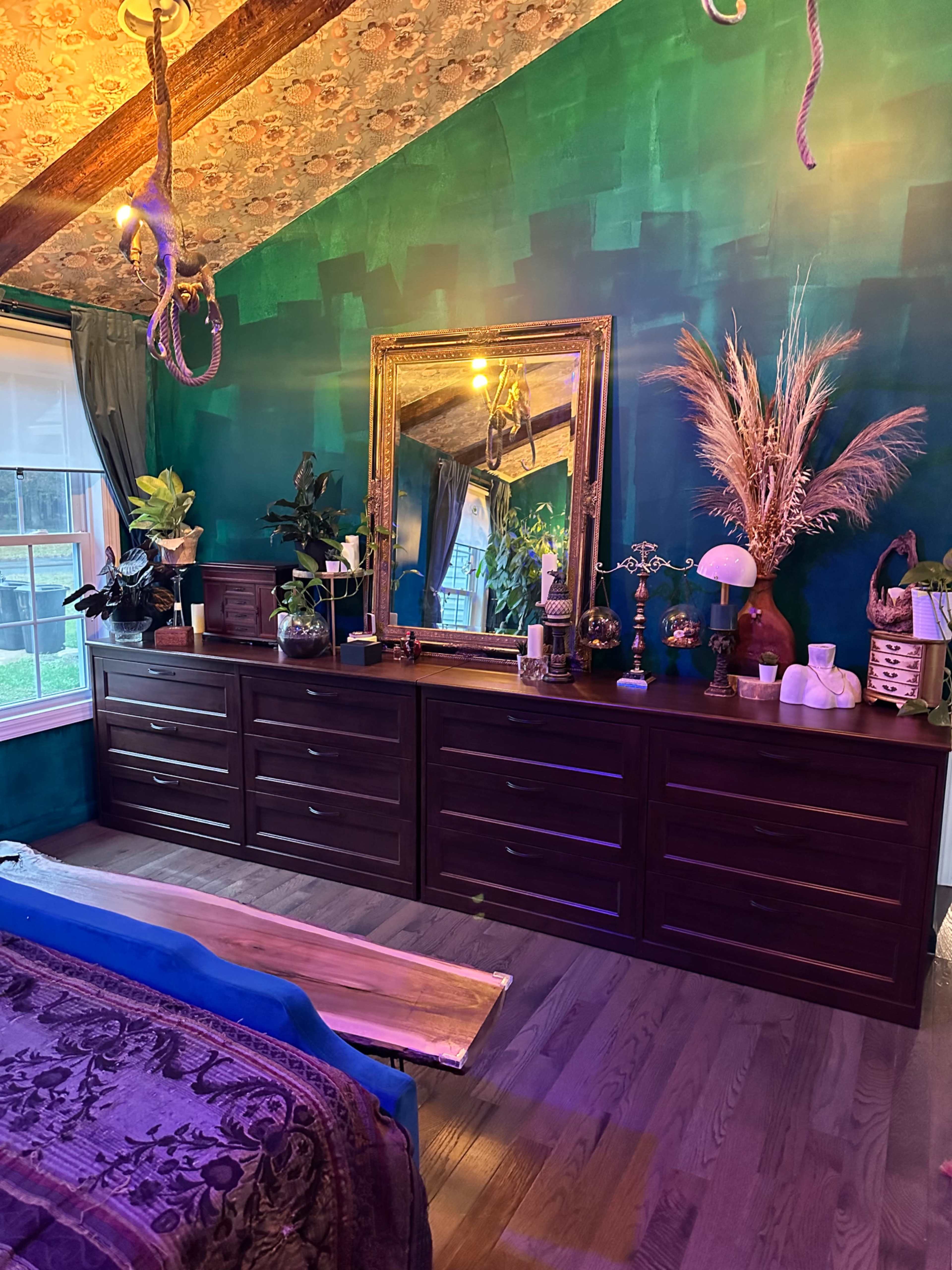 The image shows a bedroom with a wooden dresser, a large mirror above it, and various decorative plants and objects arranged on and around the dresser against a green-blue painted wall.
