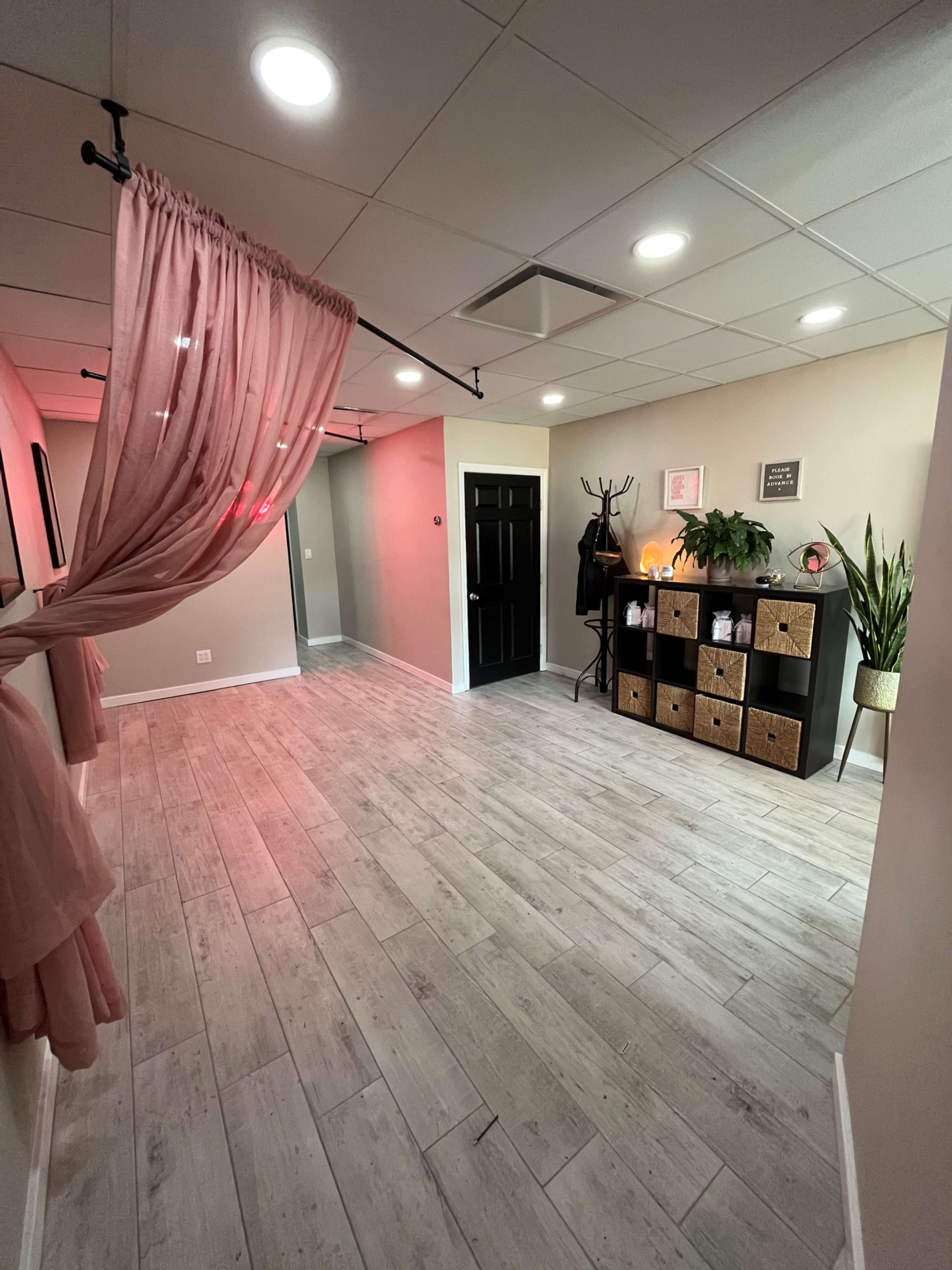 A spacious, well-lit hallway featuring a pink curtain, a black door, a shelf unit with decorative boxes, and potted plants.