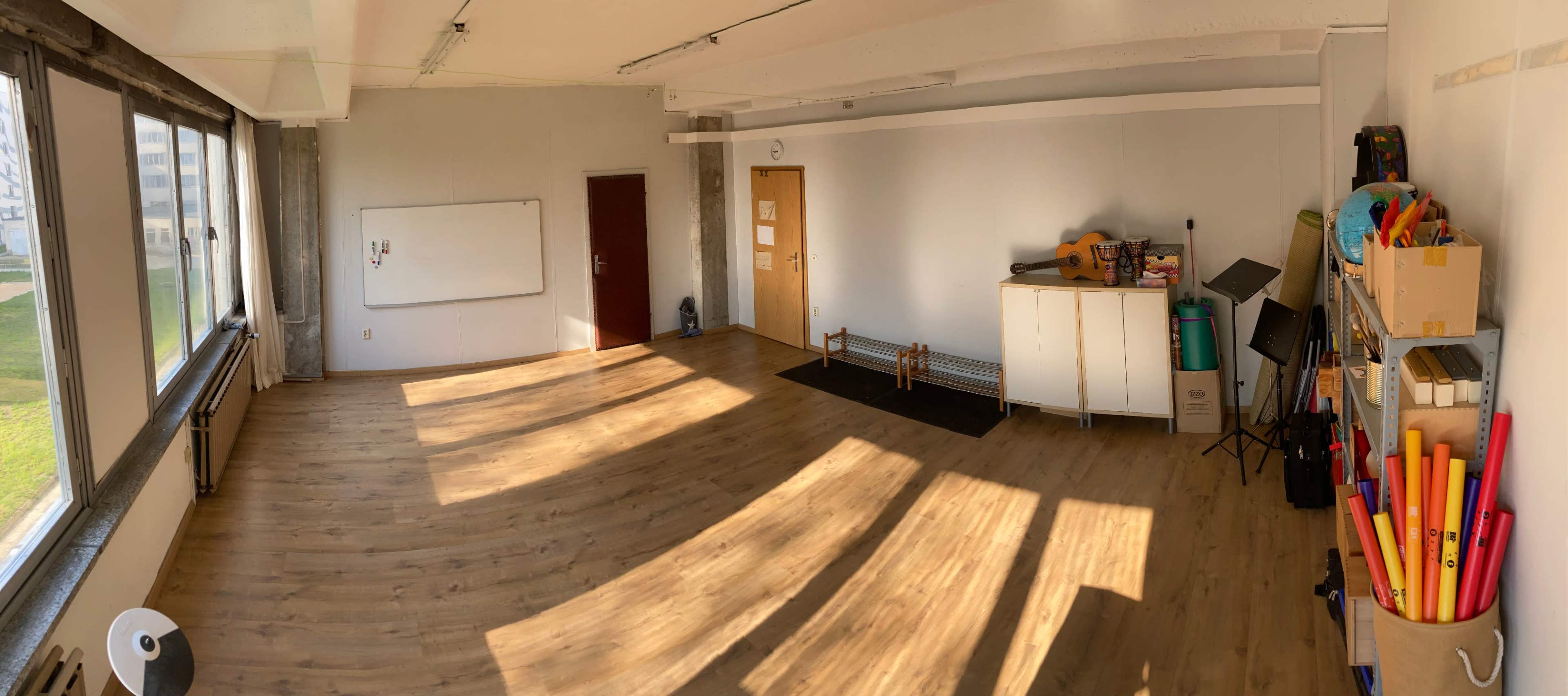 A bright, spacious classroom with wooden flooring, a whiteboard, and various teaching materials organized on shelves.