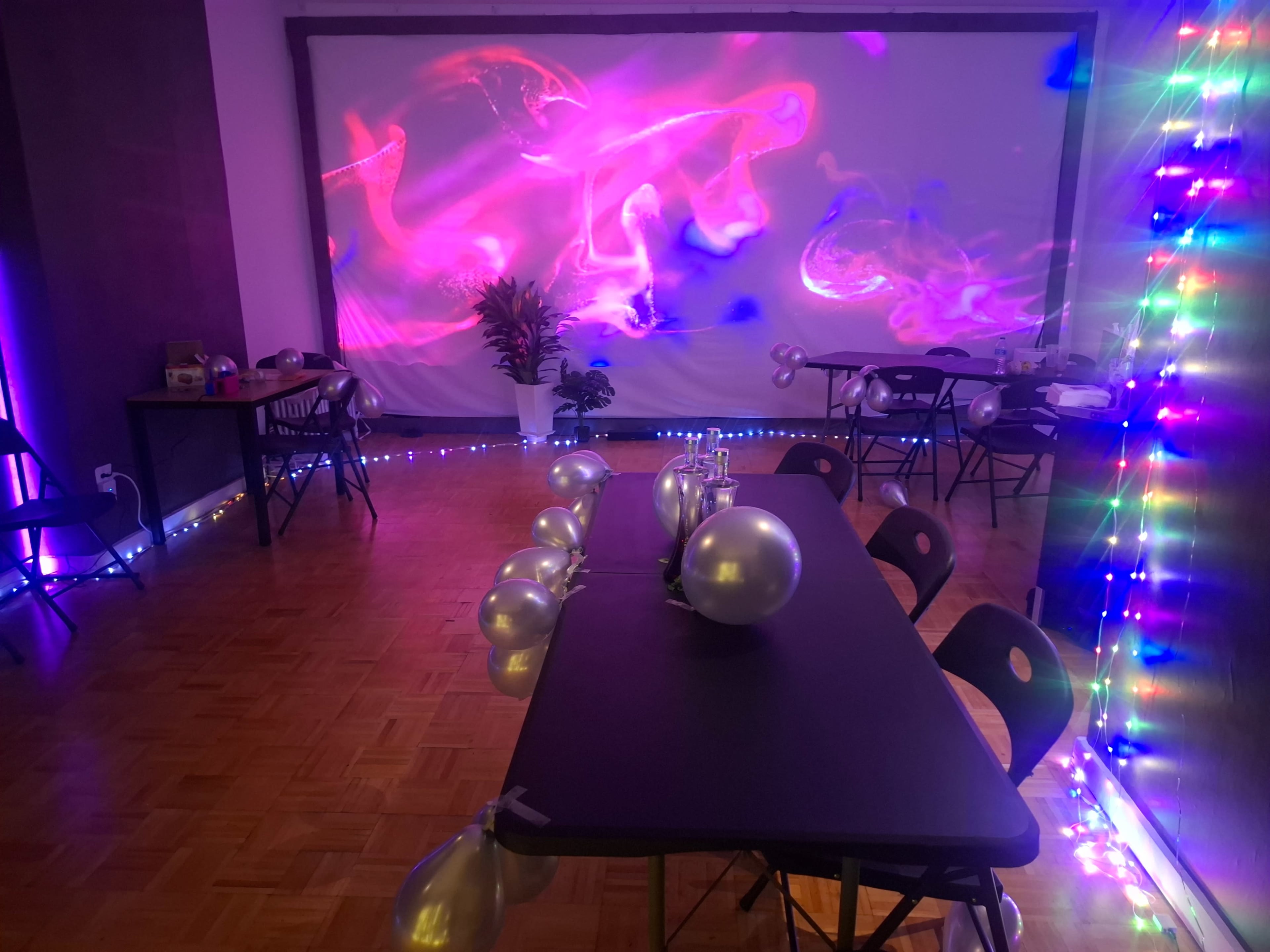 A decorated room features tables with balloons, colorful lighting, and a wall projection creating an artistic backdrop.
