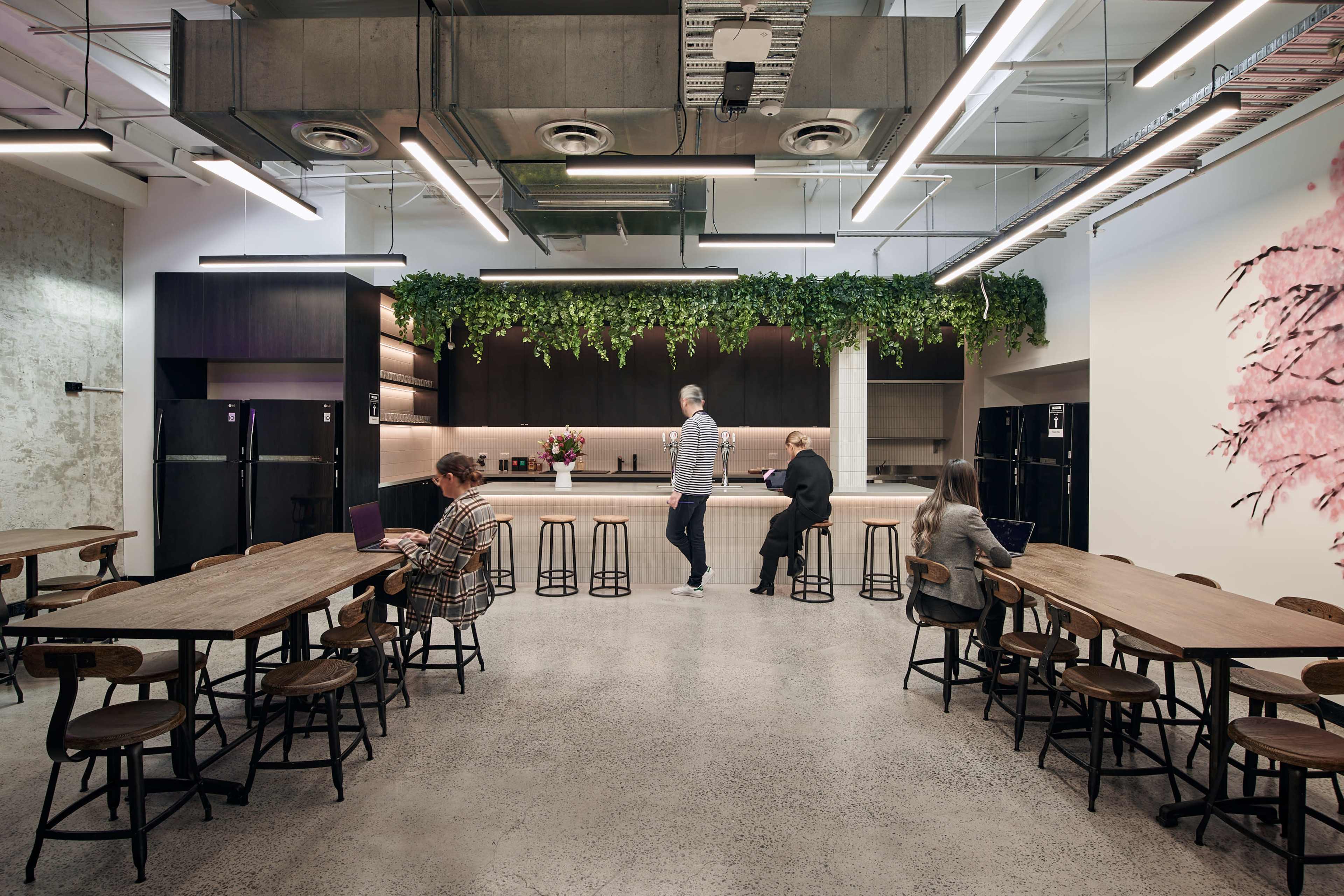 The image shows a modern kitchen and dining area with long wooden tables, black refrigerators, and decorative plants hanging from the ceiling.