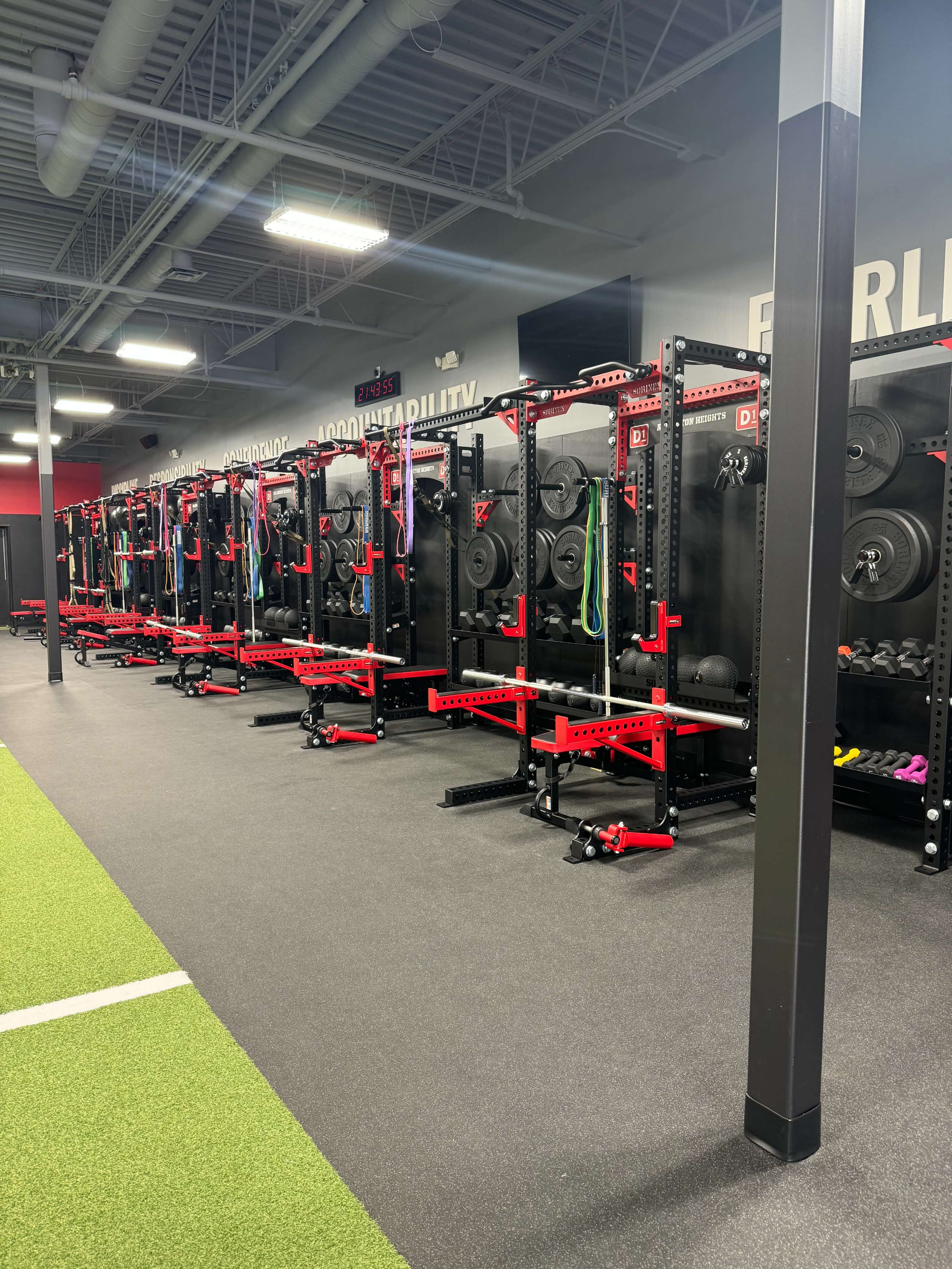 The image shows a fitness gym interior with multiple weightlifting racks lined up against a wall, equipped with various weight plates and gym accessories.