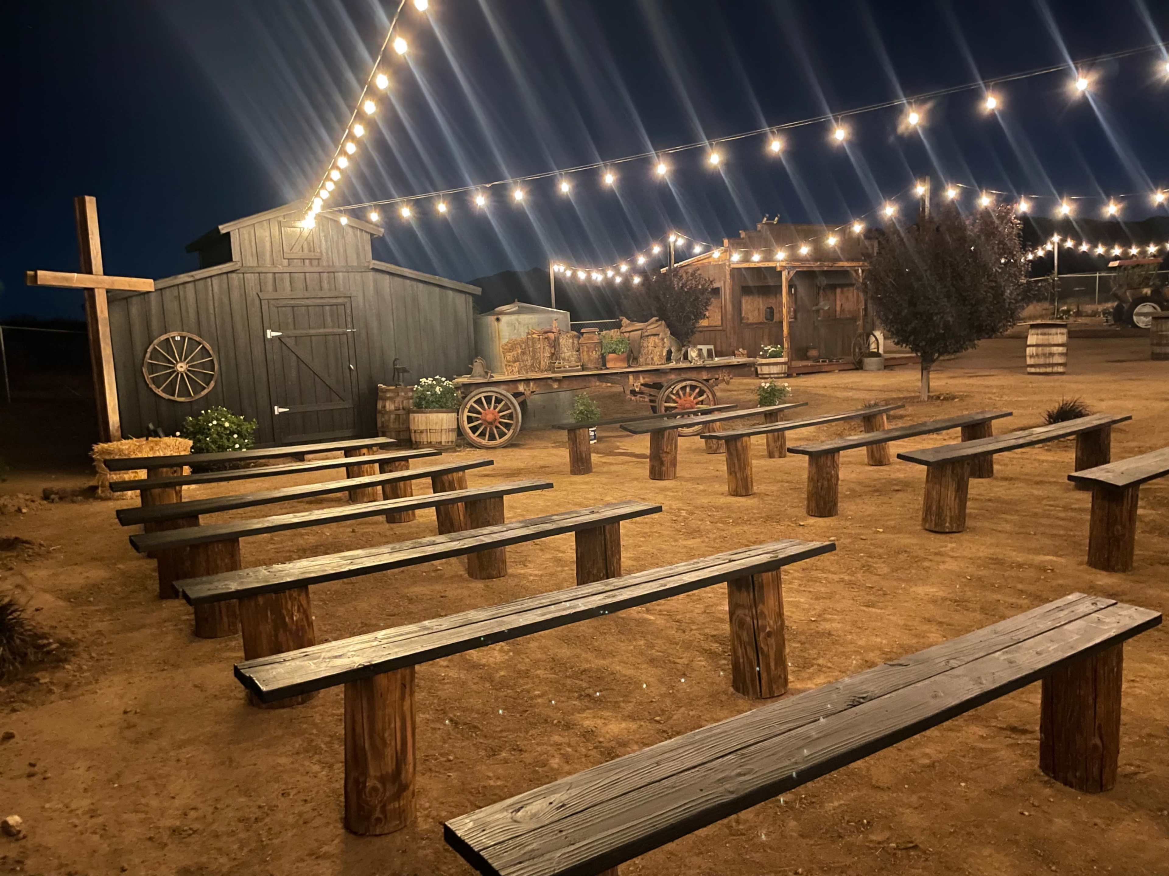 A rustic outdoor venue features wooden benches arranged in rows under string lights, with a barn and wagon in the background.
