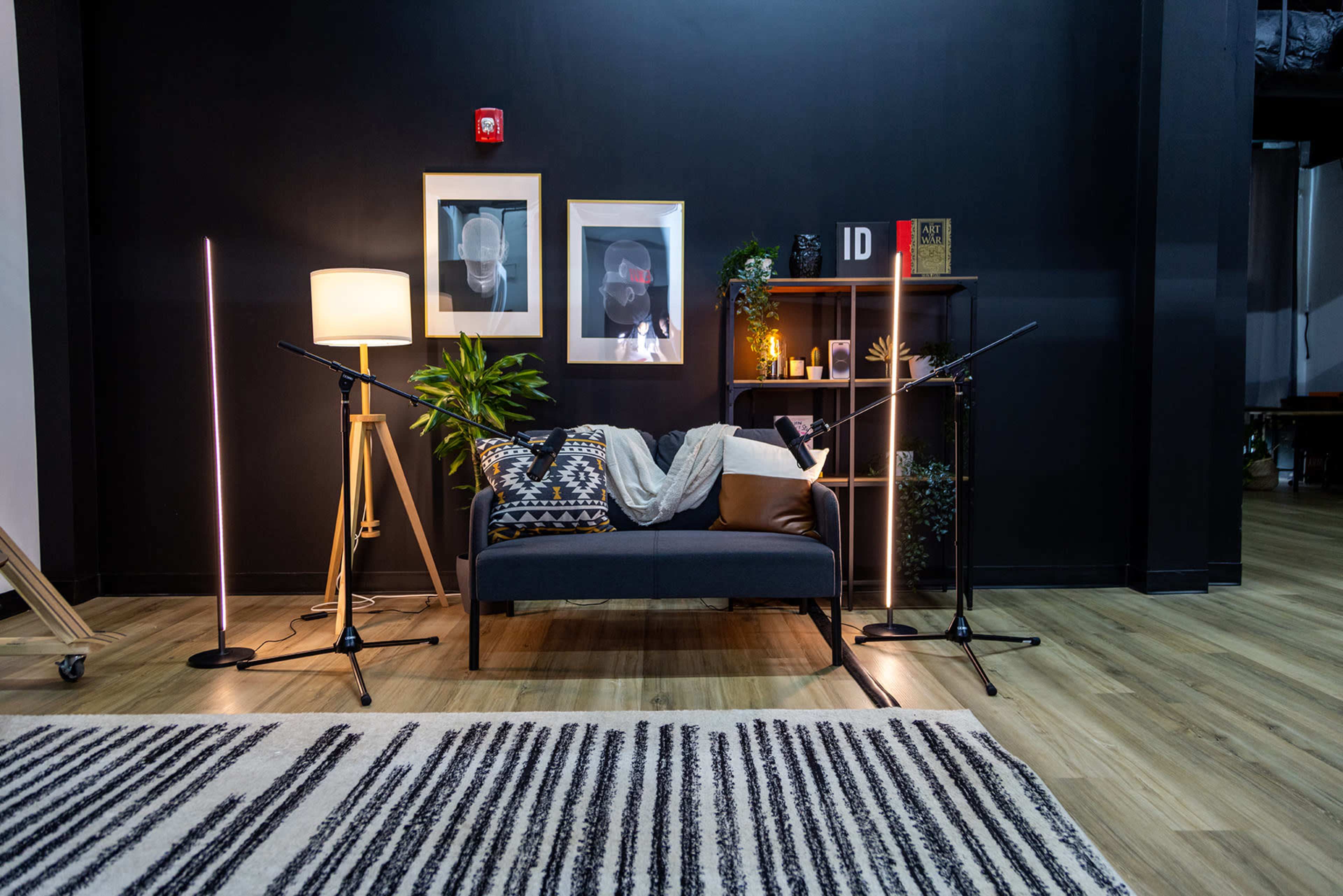 A cozy studio setting with a black wall, a sofa adorned with a blanket and pillows, and two microphone stands positioned on either side, surrounded by decorative plants and a lamp.