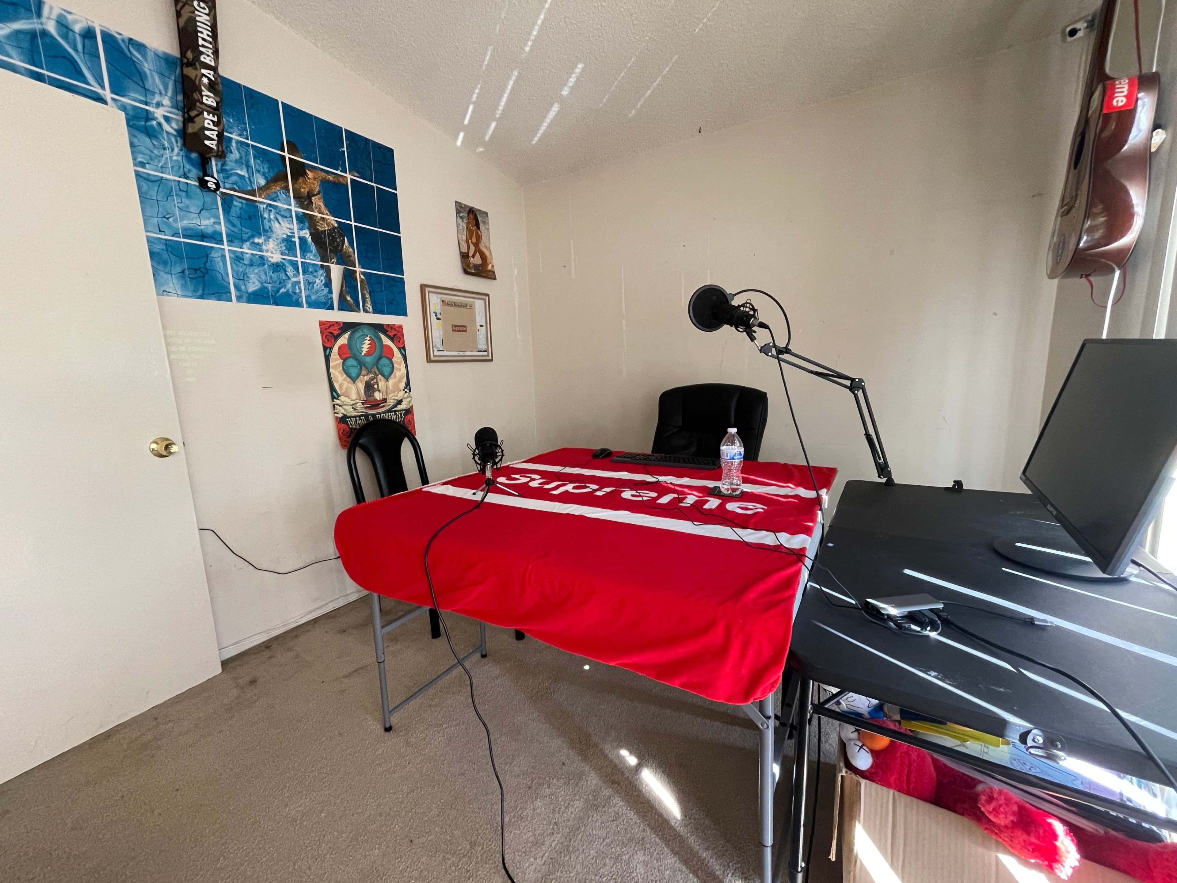 A small, sparsely furnished room with a table covered by a red Supreme blanket, two microphones set up on either side, a chair, and a desk with a monitor.
