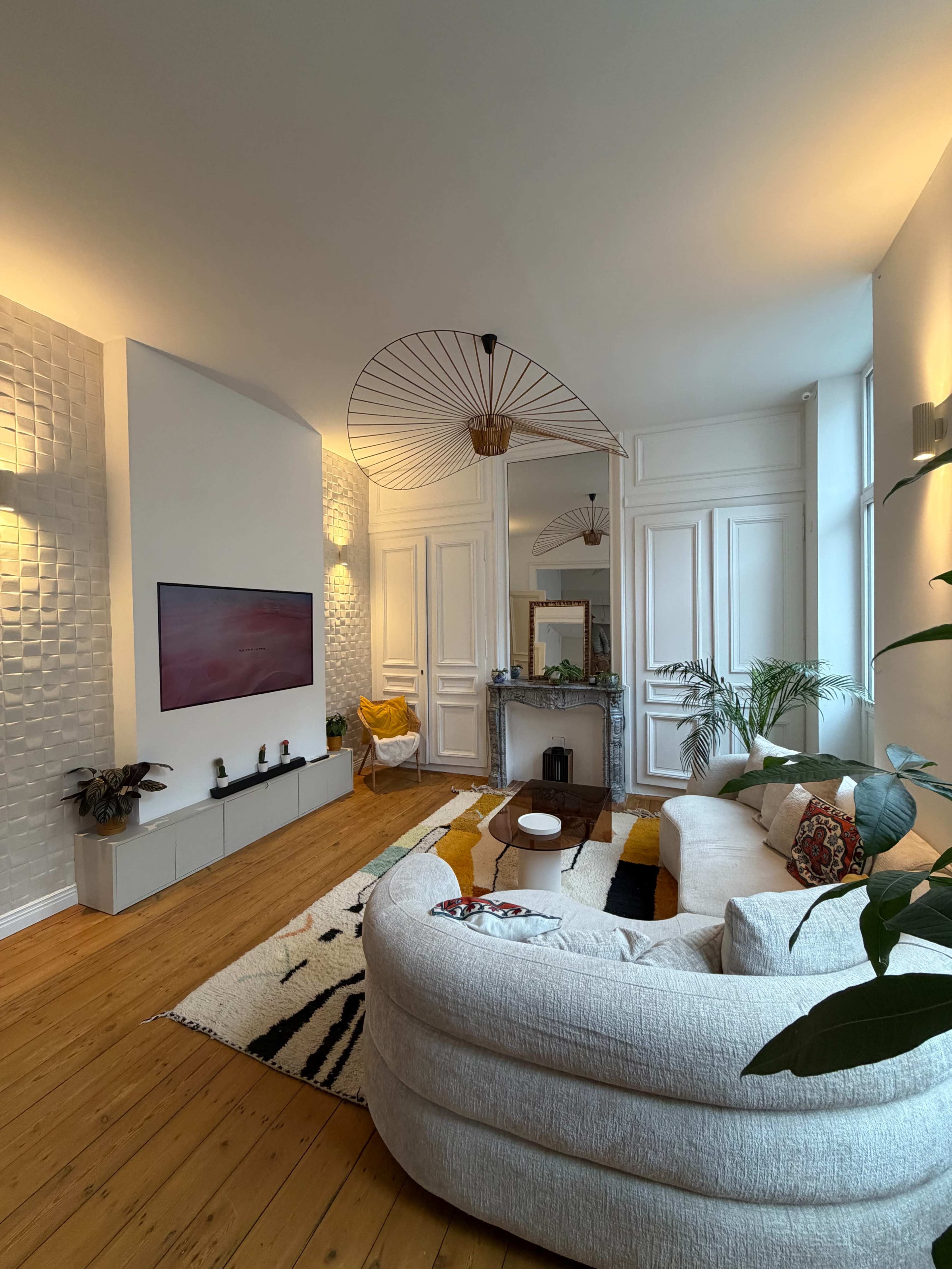 The image shows a modern living room featuring a circular white sofa, a wall-mounted TV, and decorative plants, with light-colored walls and hardwood flooring.