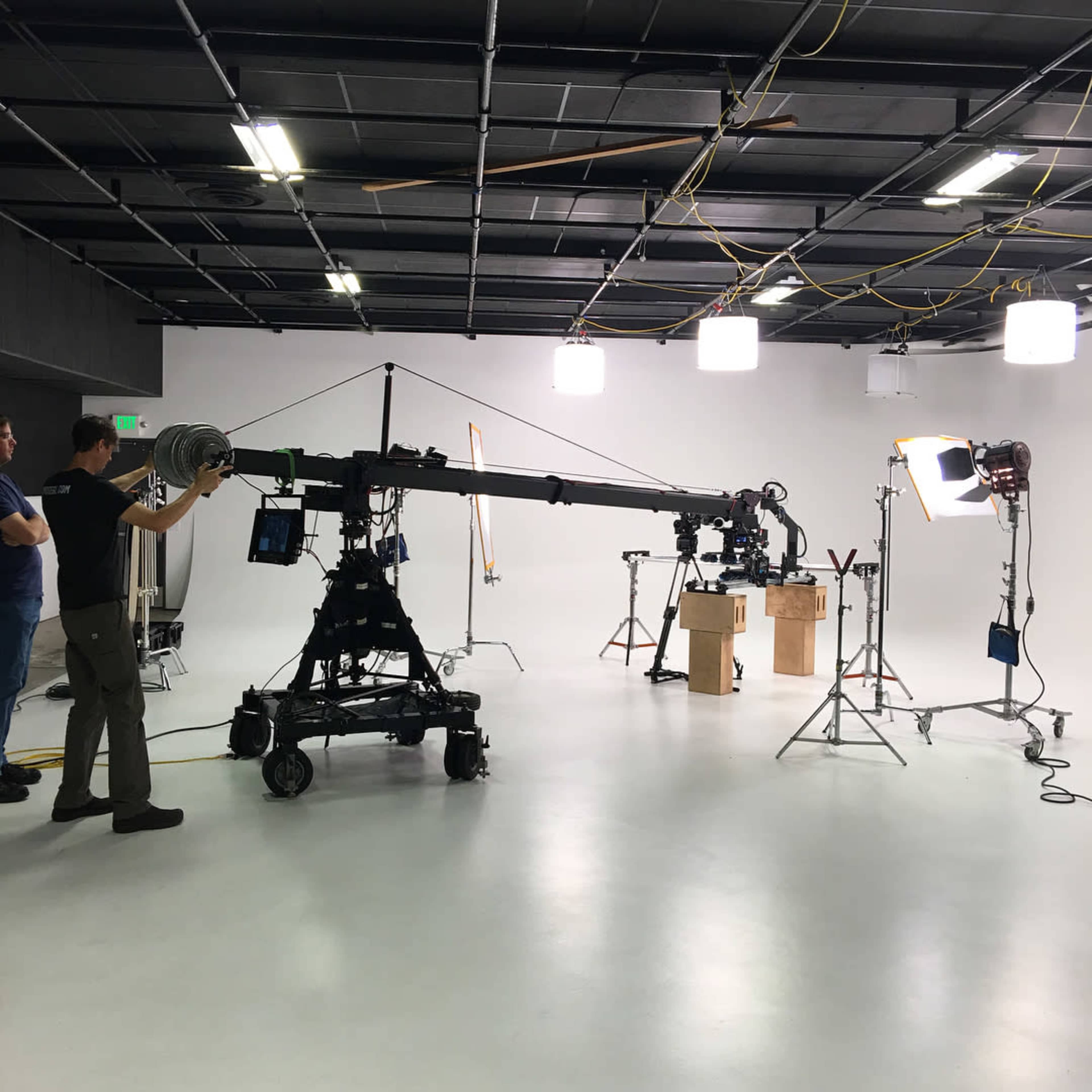 A film crew operates a camera crane in a well-lit studio set with various lighting equipment and props.