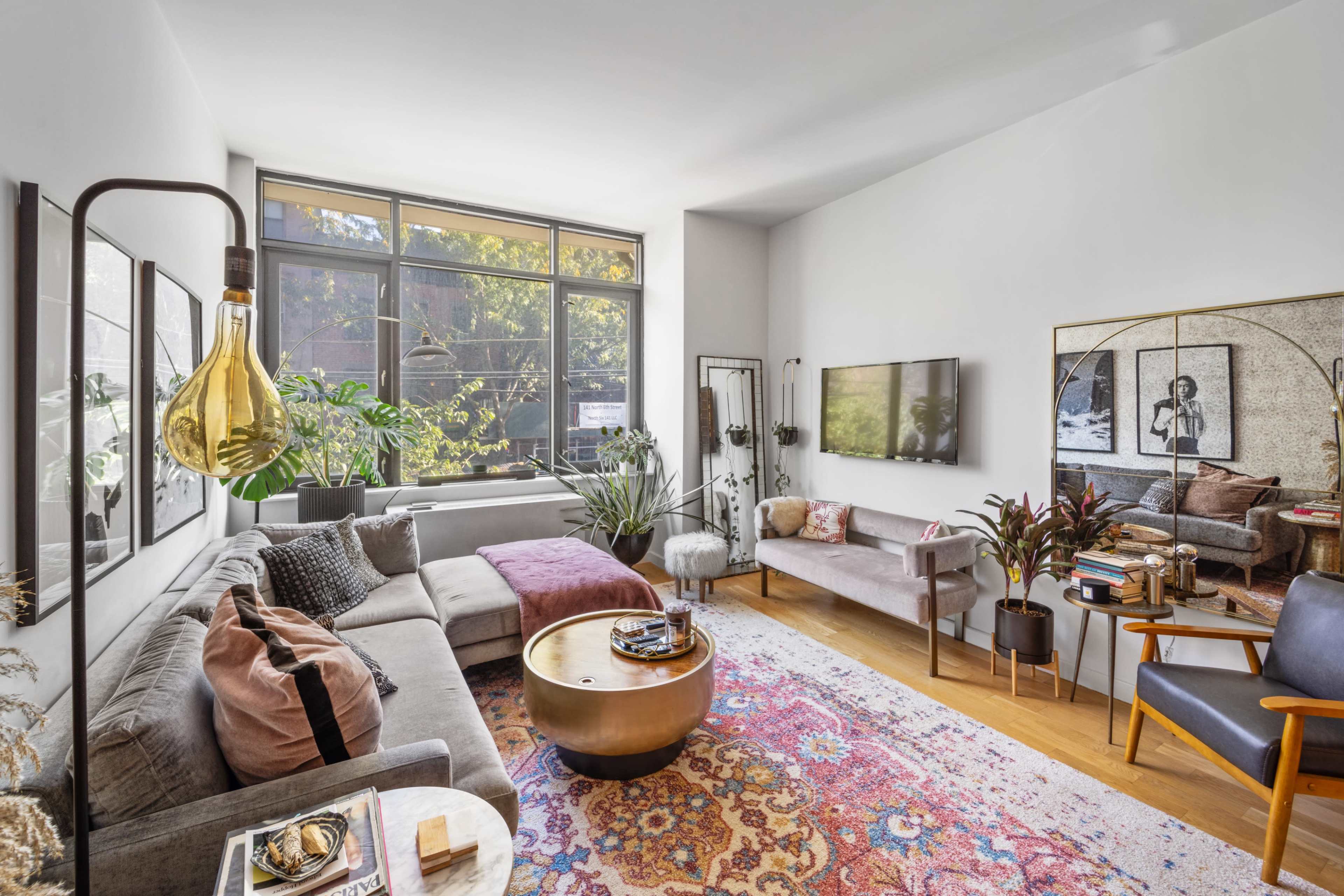 A modern living room features a gray sectional sofa, a large window with plants outside, a round coffee table, and a decorative rug.