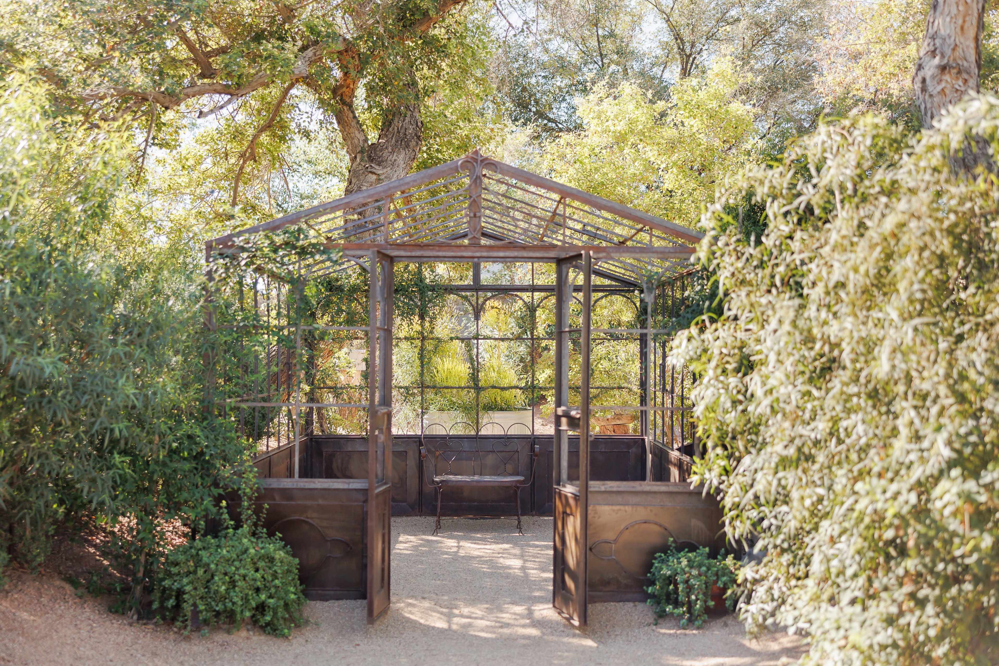 The image shows a rustic, open-air structure with a metal frame and glass panels surrounded by greenery in a garden setting.