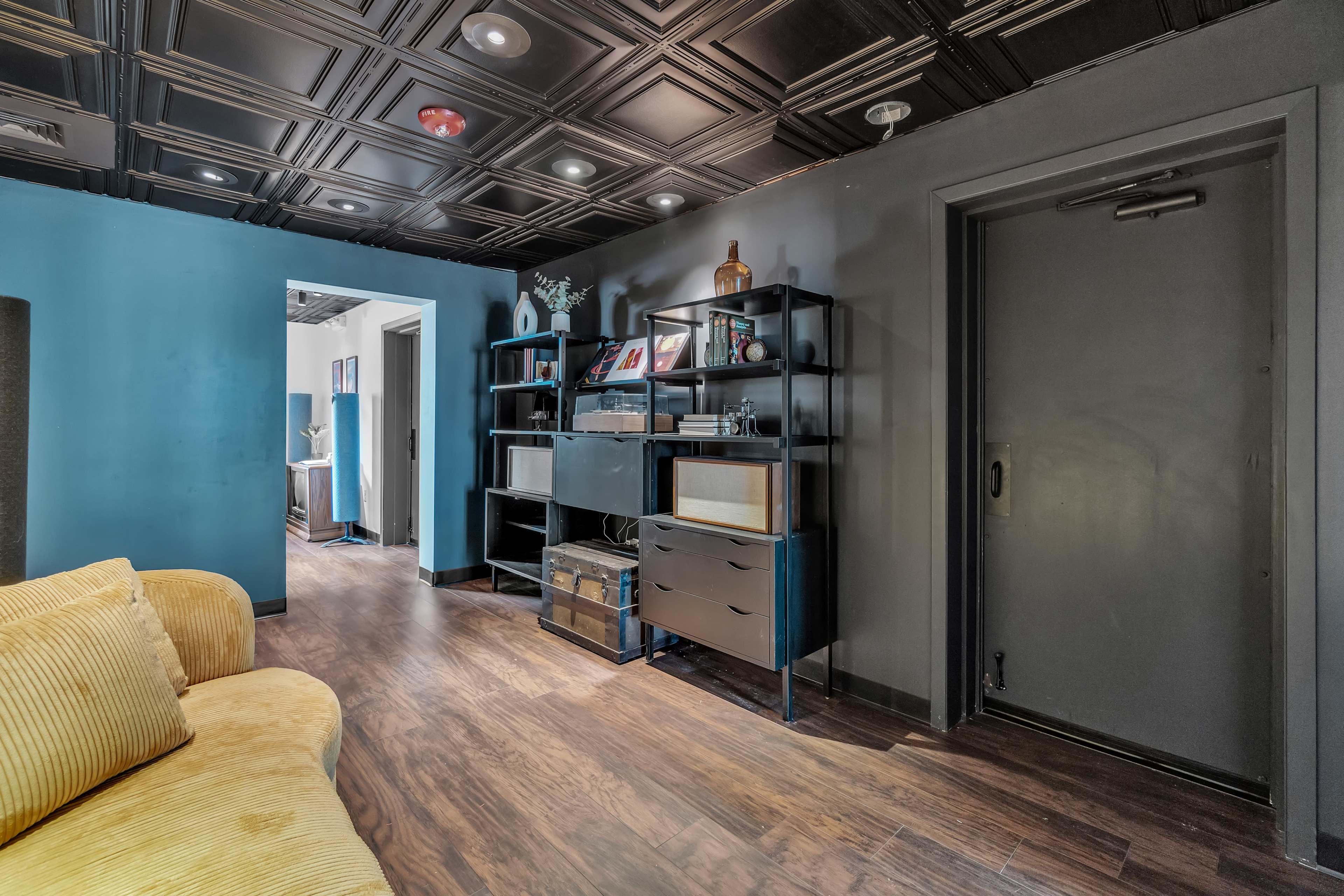 The image shows a modern interior space with a dark blue wall, a black shelving unit, and a yellow couch, illuminated by overhead lights.