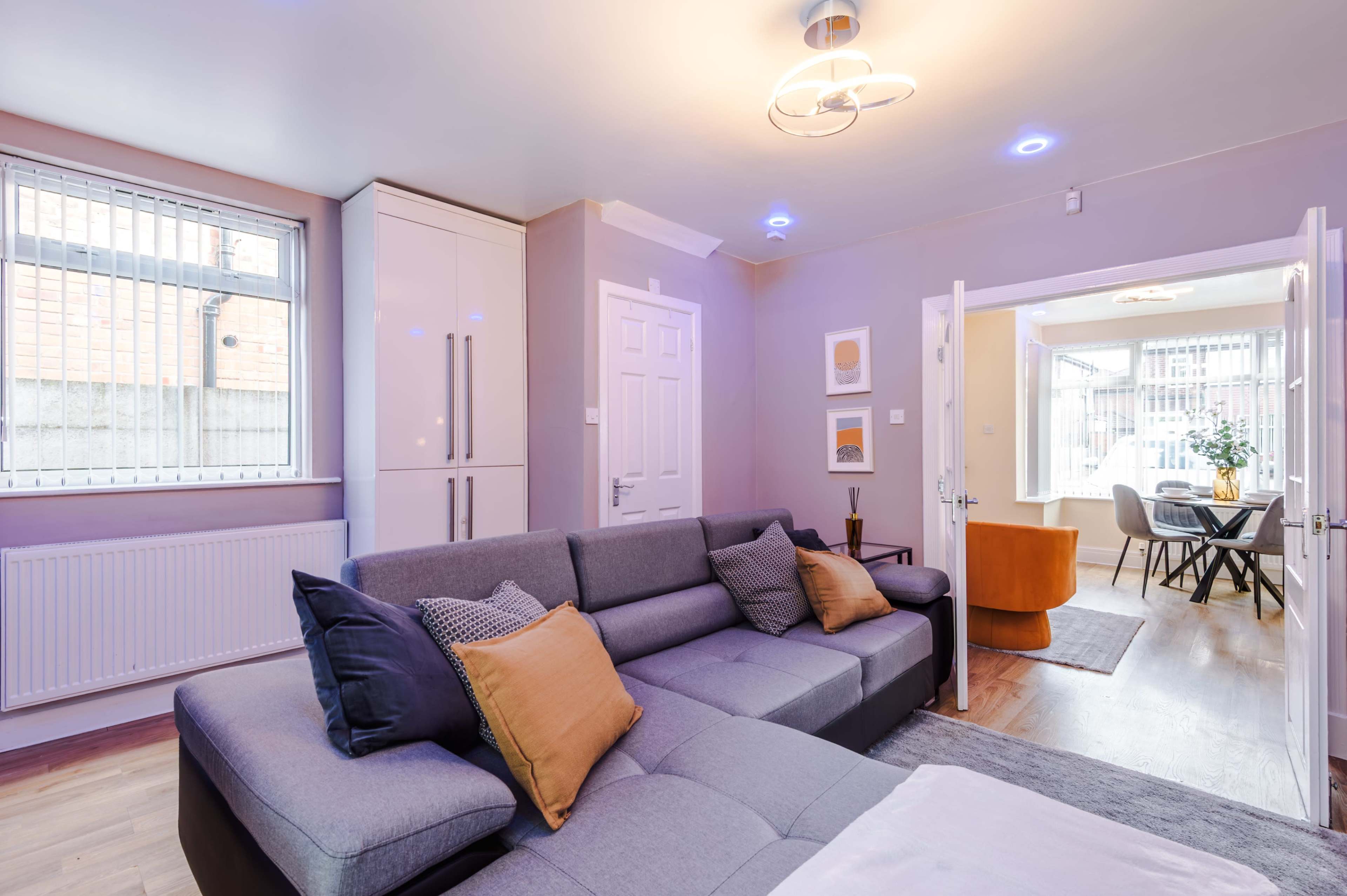 The image shows a modern living room featuring a gray sectional sofa with decorative pillows, light-colored walls, and an adjacent dining area visible through open doors.