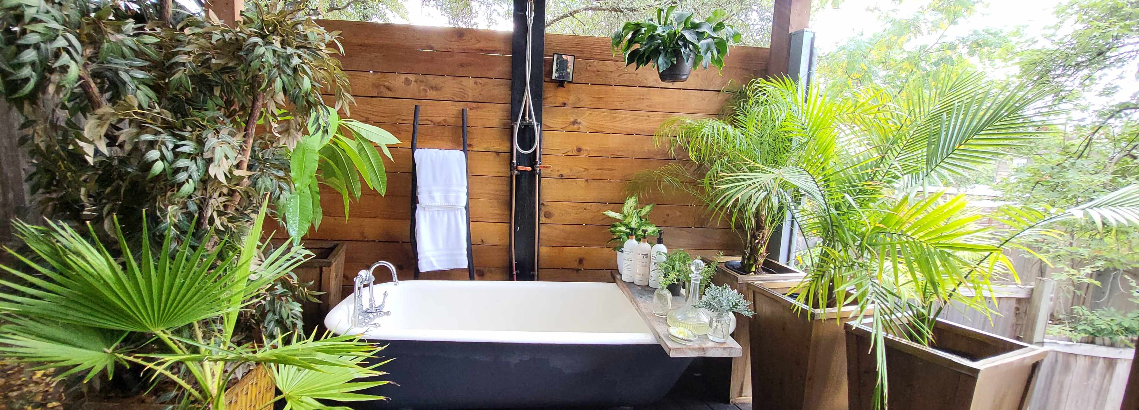 The image shows an outdoor bathroom with a freestanding bathtub surrounded by various plants and wooden walls.