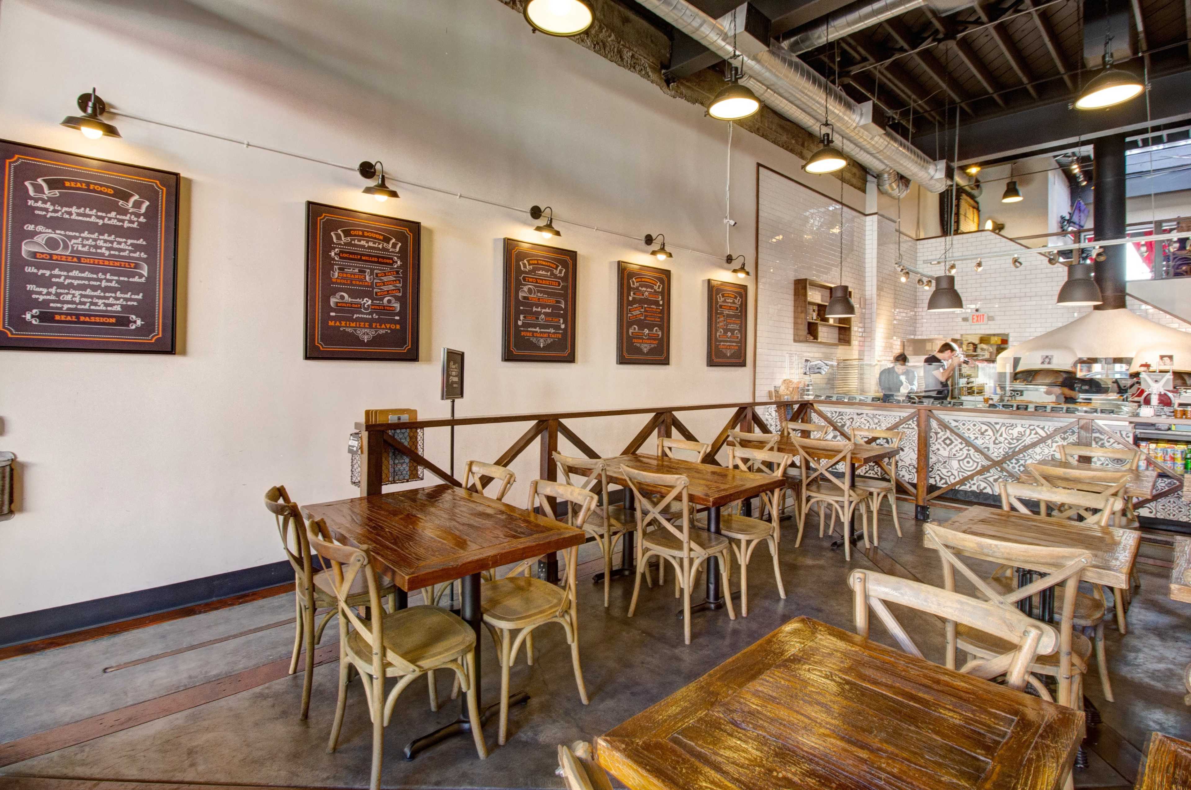A modern café with wooden tables and chairs, exposed industrial lighting, and a menu display on the wall.