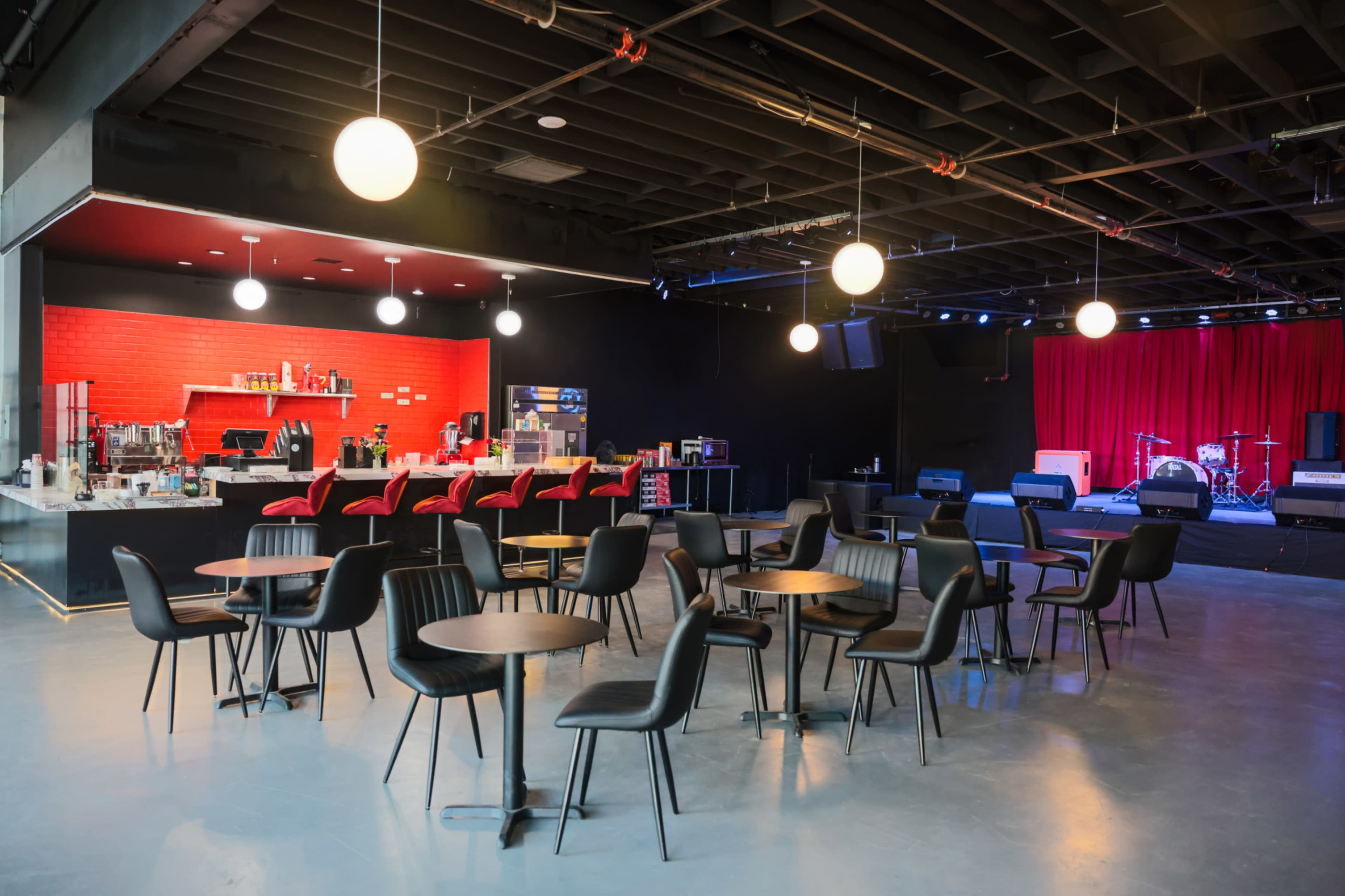 The image shows an empty performance space with round tables and black chairs, a red-accented bar area in the background, and a stage set for a live event.