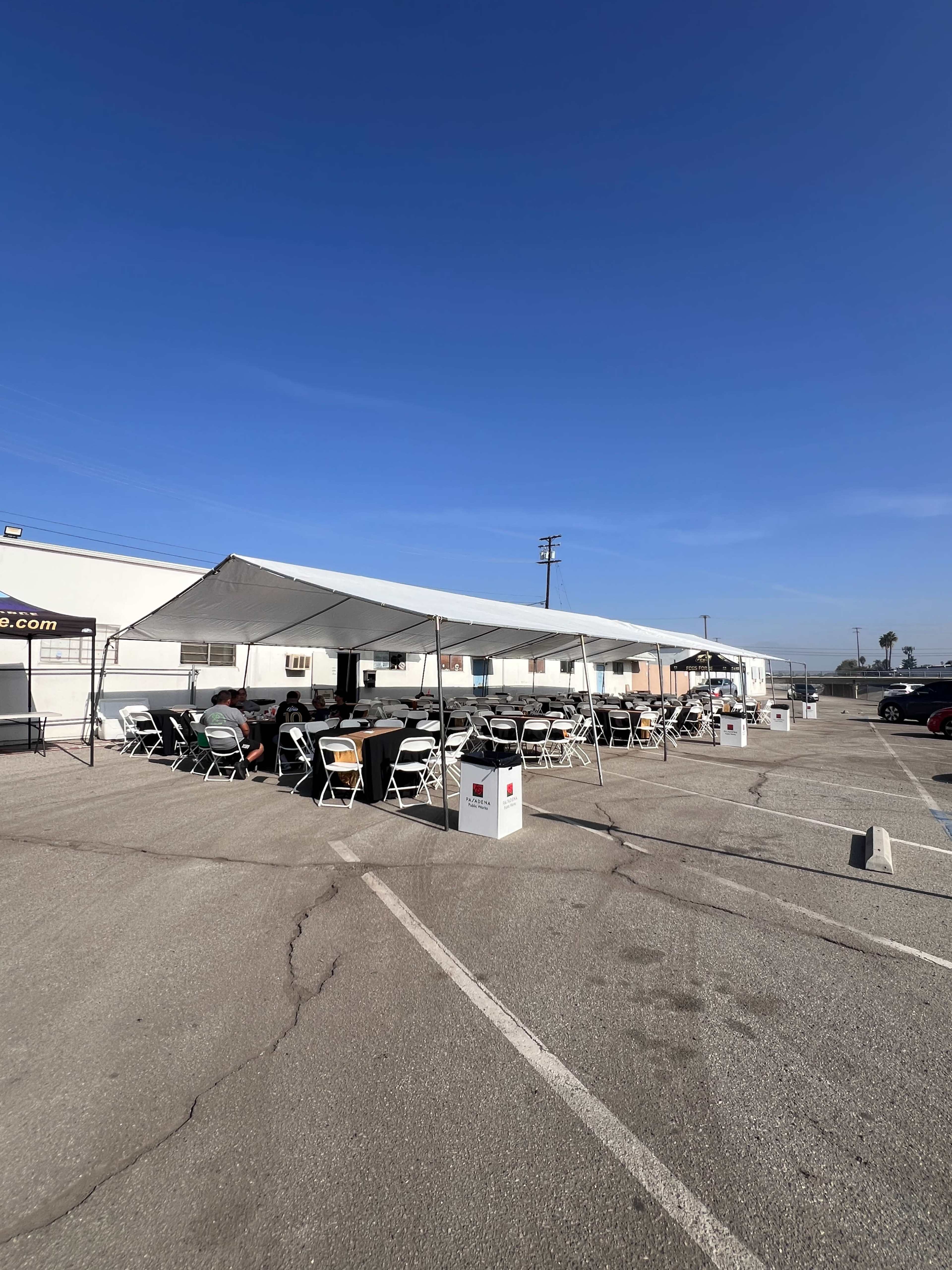A large tent with rows of tables and chairs is set up in a parking lot under a clear blue sky.