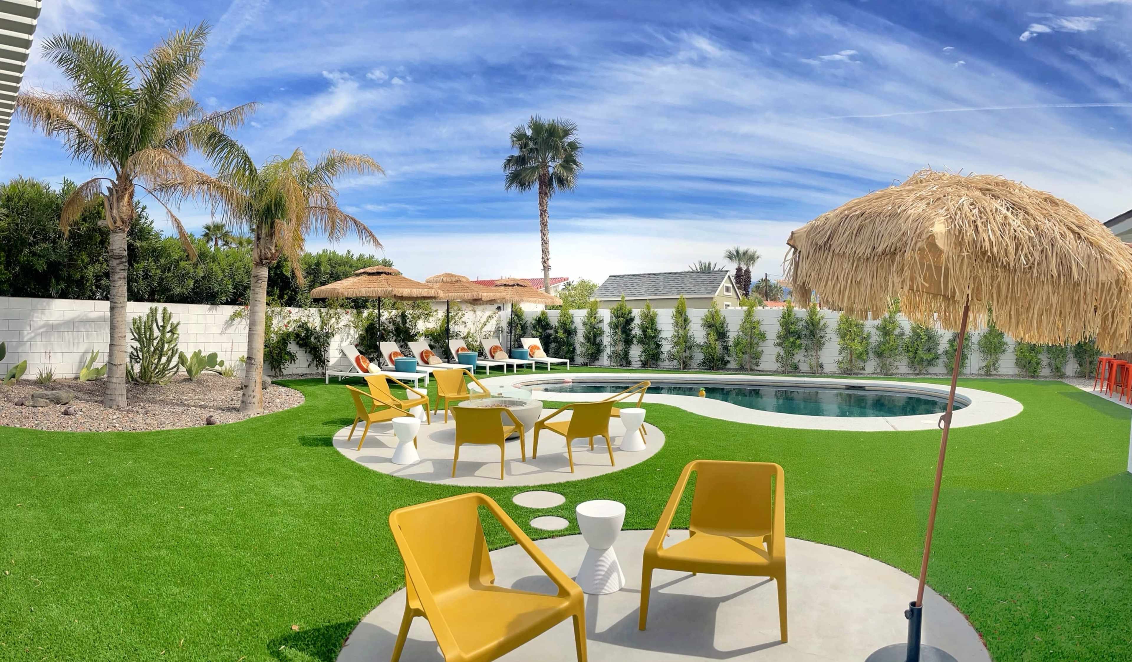 A backyard pool area features a circular swimming pool surrounded by lounge chairs, a thatched umbrella, and palm trees under a blue sky.