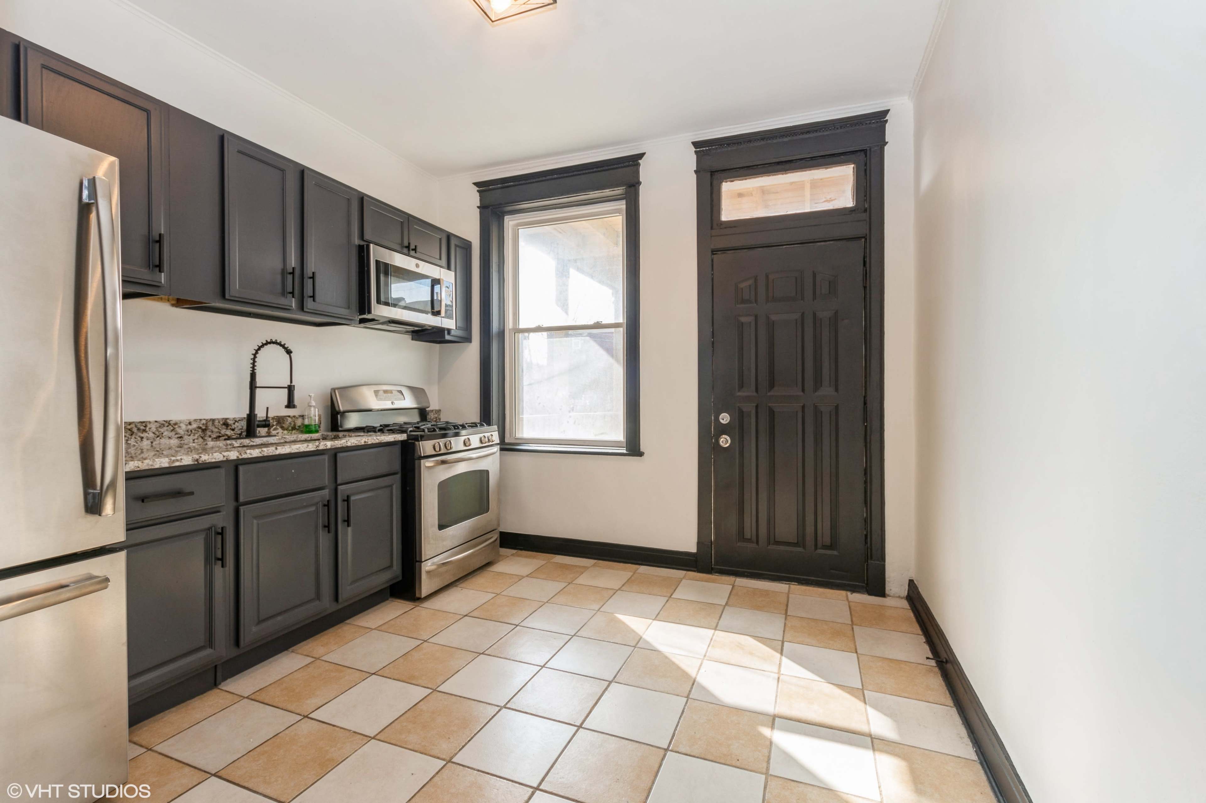 The image shows a kitchen with dark cabinets, a stainless steel refrigerator, a stove, and a tiled floor.