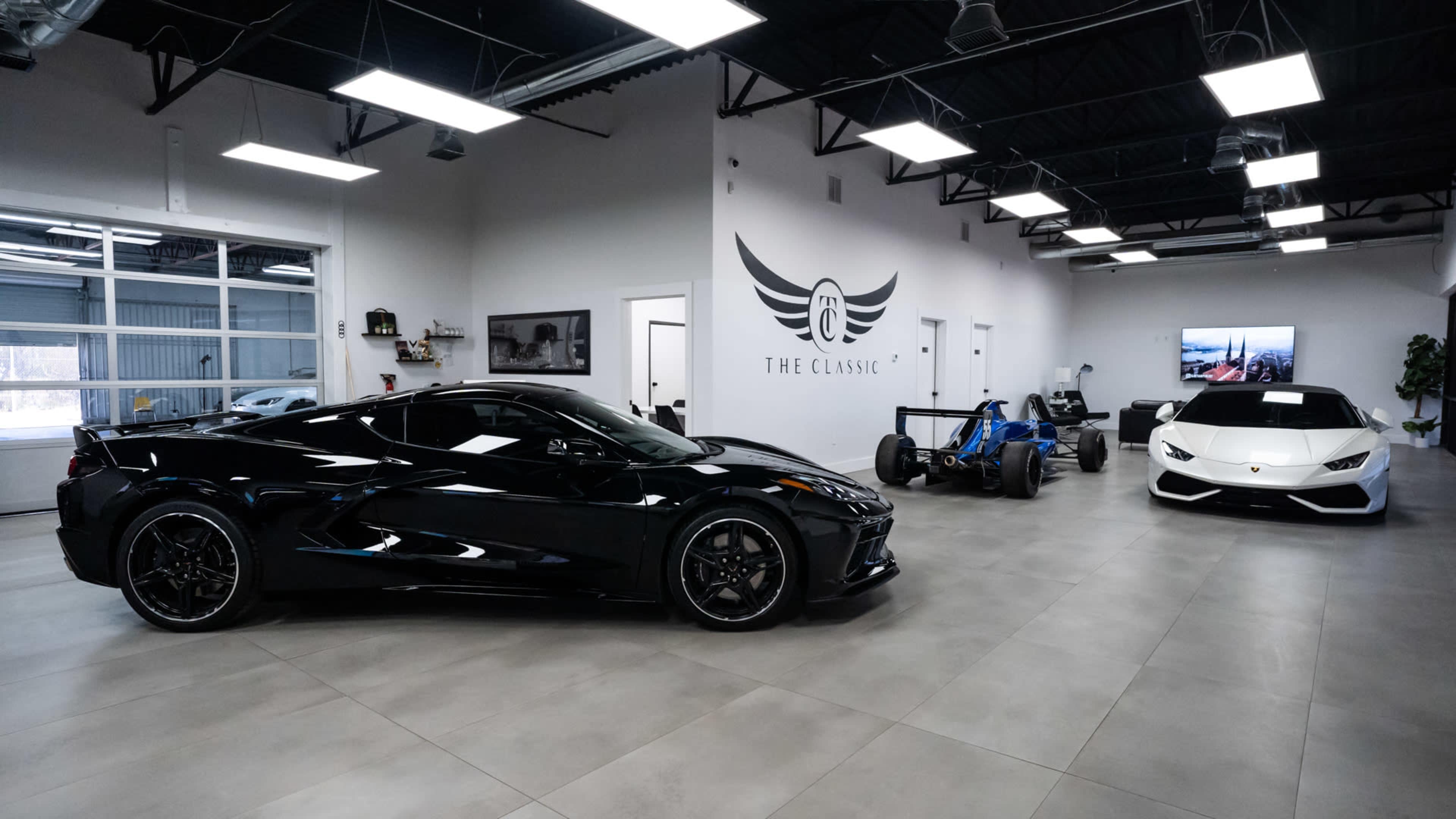A sleek car showroom featuring a black sports car, a blue racing car, and a white luxury vehicle, all displayed under bright lights in a modern, clean space.