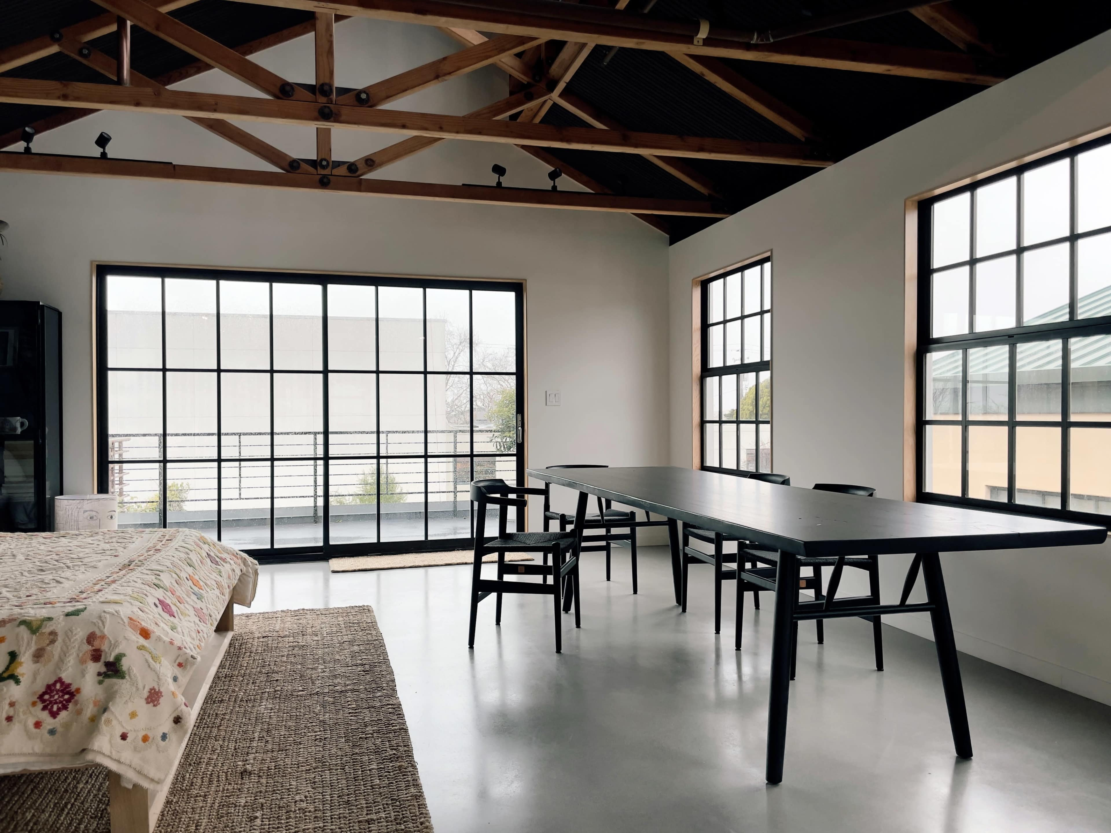 A spacious room with a large dining table and multiple windows allowing natural light, featuring a simple design with wooden beams.