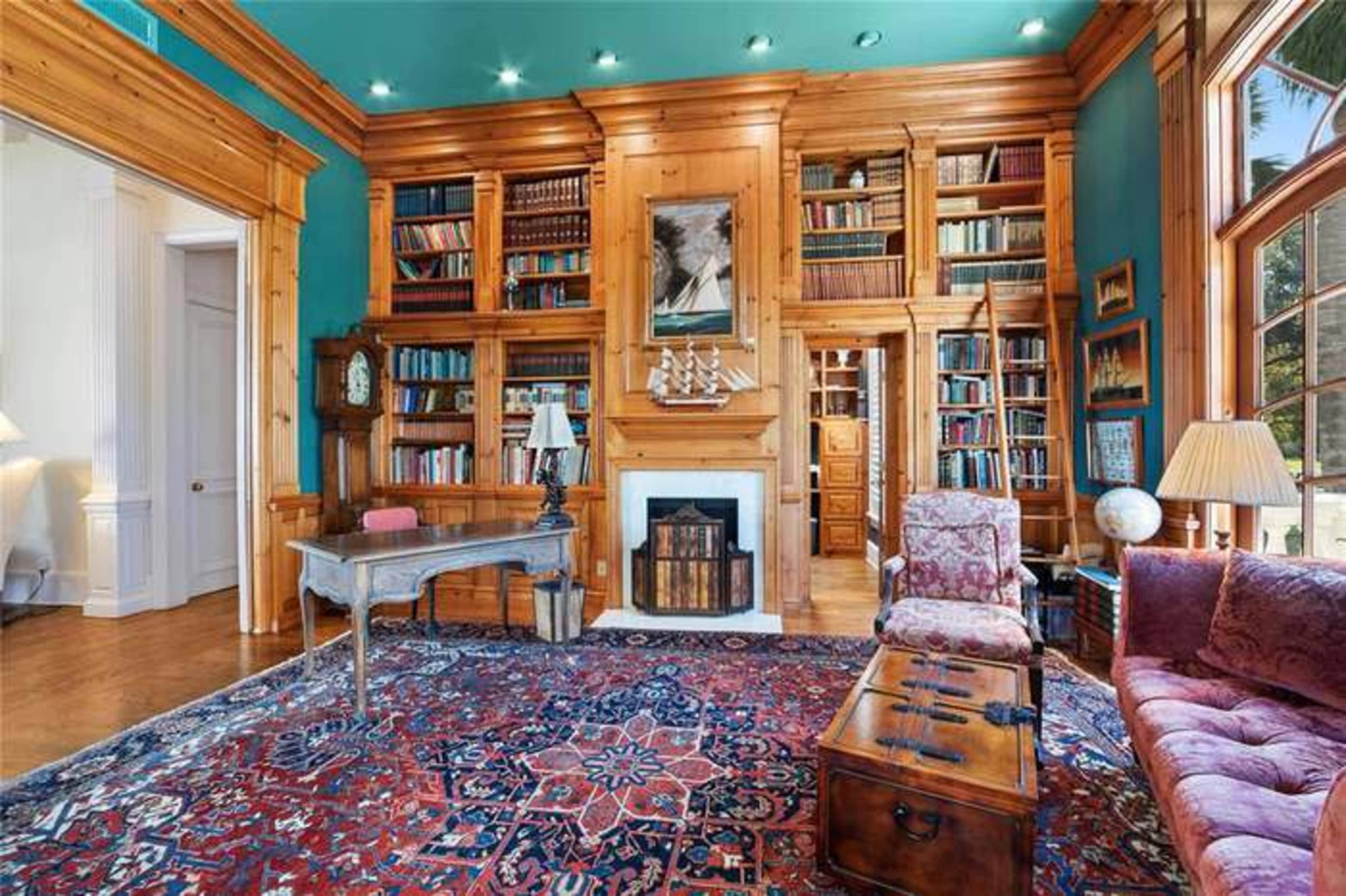 A wooden-paneled room featuring a large bookshelf, a fireplace, a patterned rug, and a seating area with an armchair and a sofa.