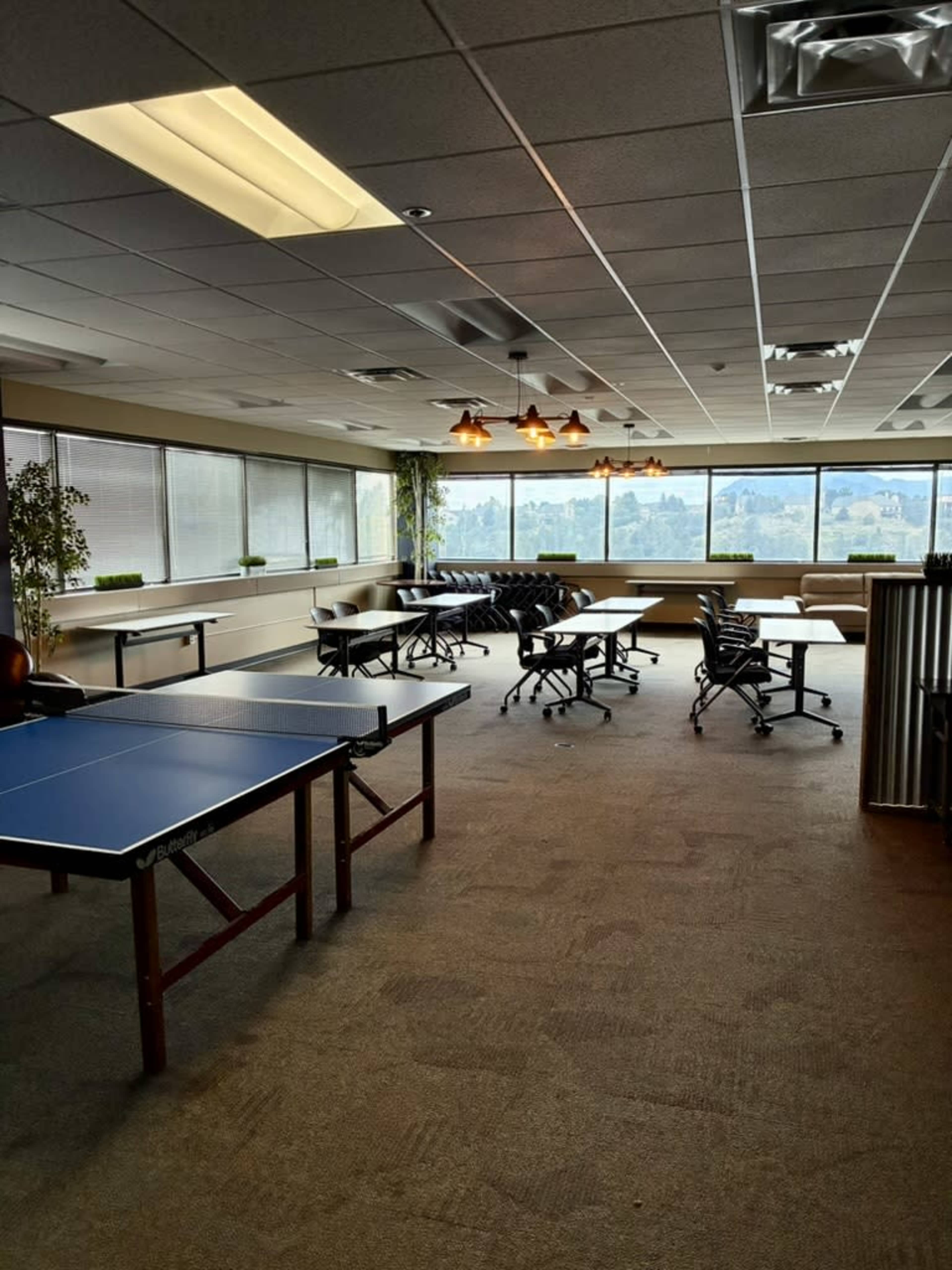 The image shows an office lounge area with a ping pong table, several chairs, and large windows offering a view of the outdoors.