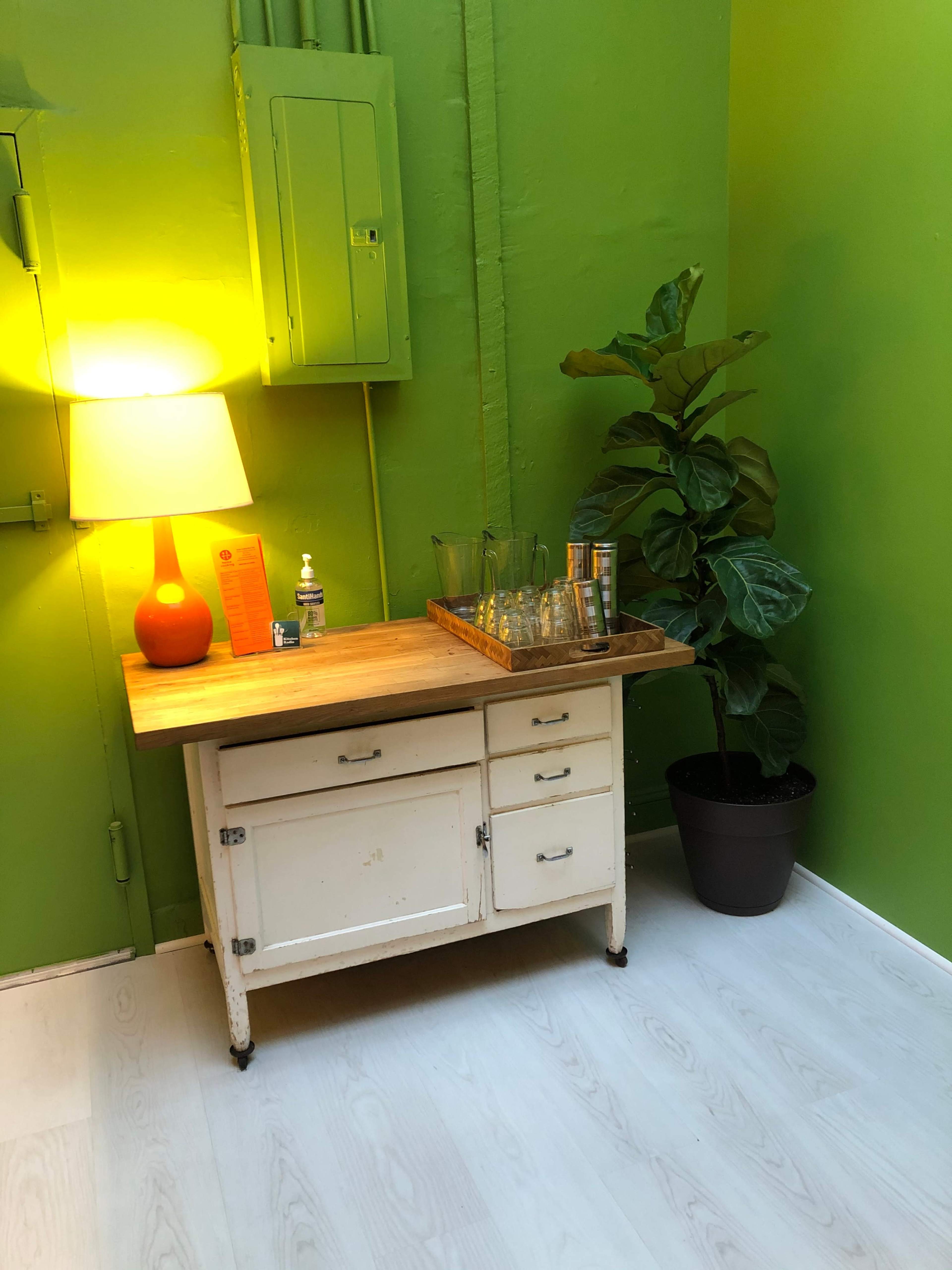 A small white side table with a wooden top holds a tray of glasses and a bottle, next to a lamp and a potted plant in a bright green room.