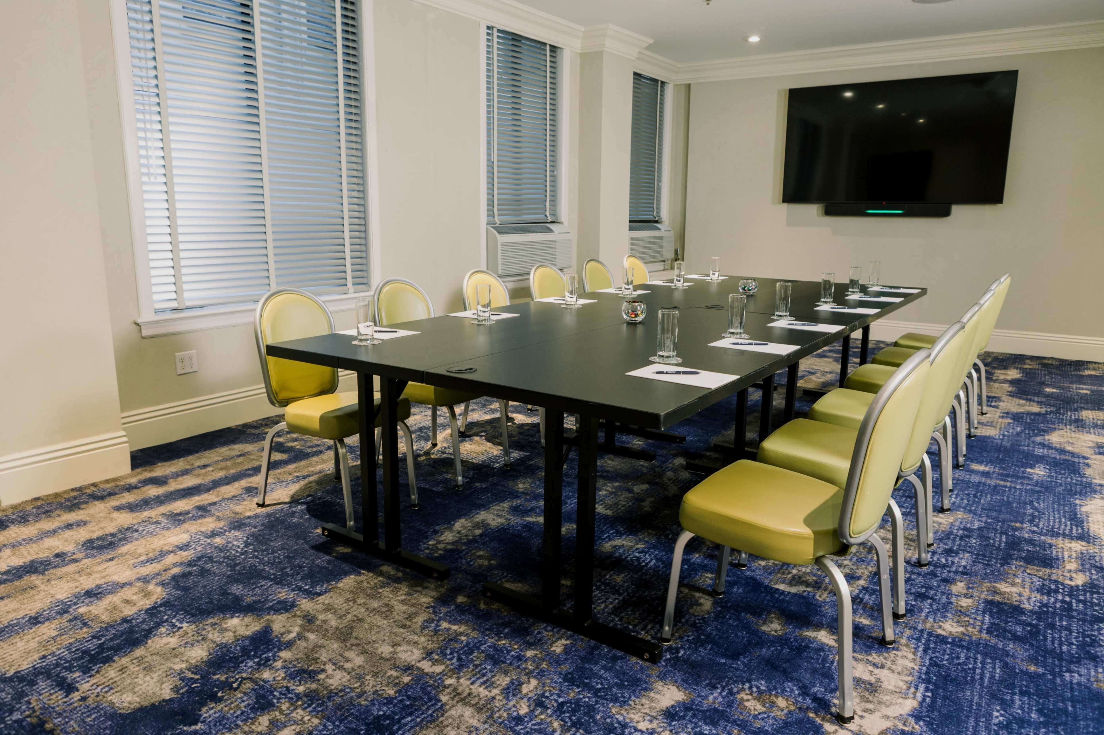 A conference room with a large black table surrounded by twelve yellow chairs and equipped with a wall-mounted screen.
