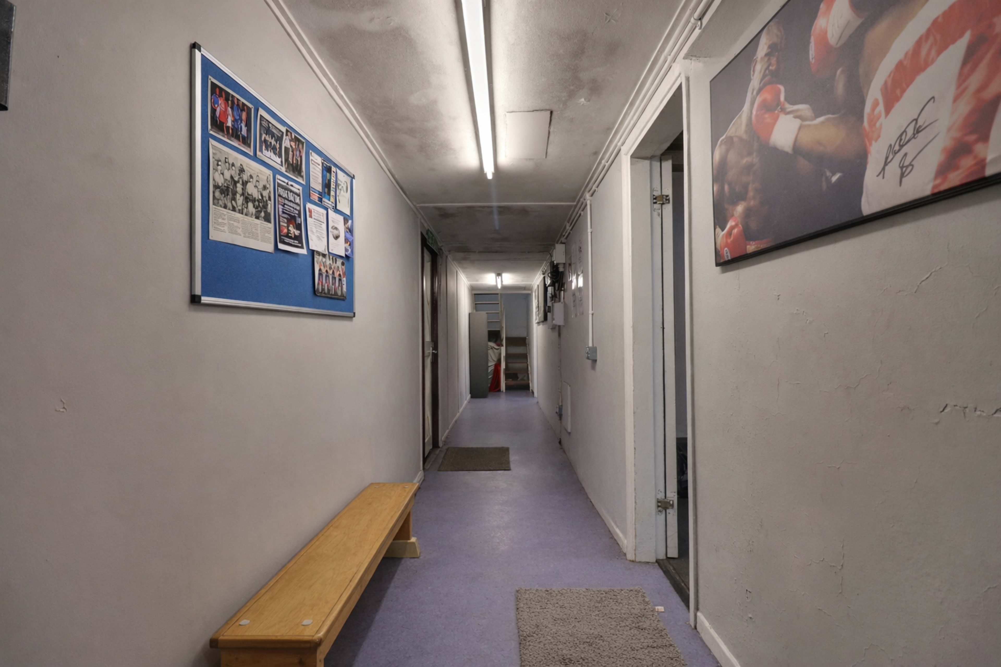 A long, narrow hallway with a wooden bench, blue bulletin board, and several doors on either side.