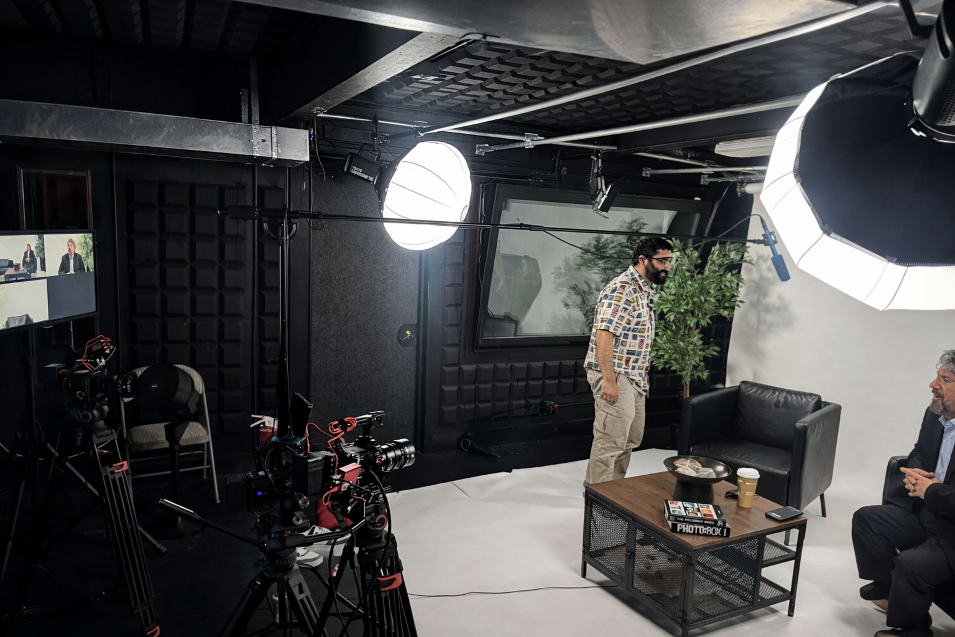 A video recording studio is set up with cameras, bright lights, and a table, where a man with glasses stands beside a chair.