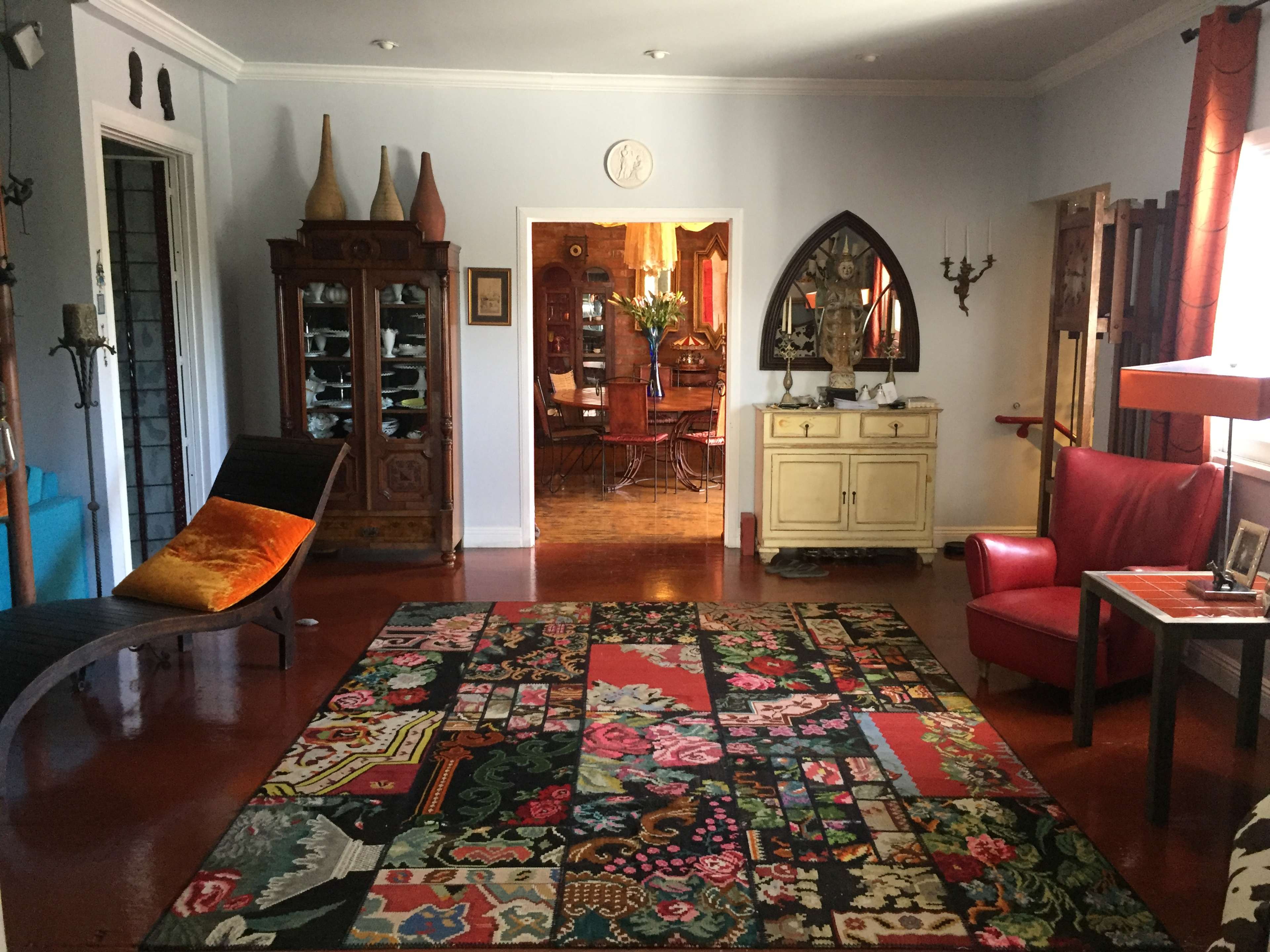 The room features a colorful rug, a red armchair, a wooden cabinet, and various decorative items, with a glimpse of a dining area in the background.