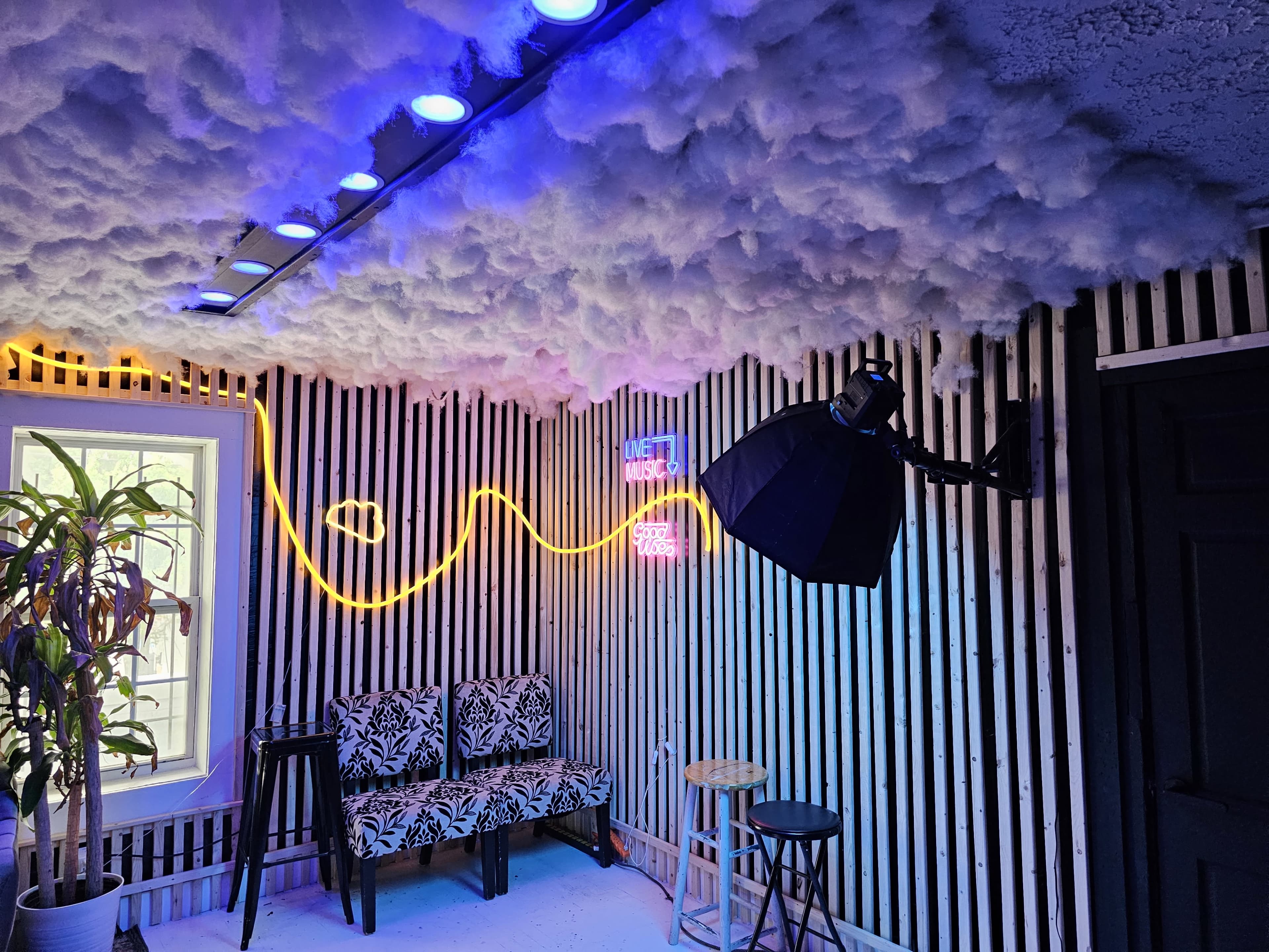 The image shows a room with striped walls, a cloud-like ceiling, neon lights, a photography softbox, and two chairs.
