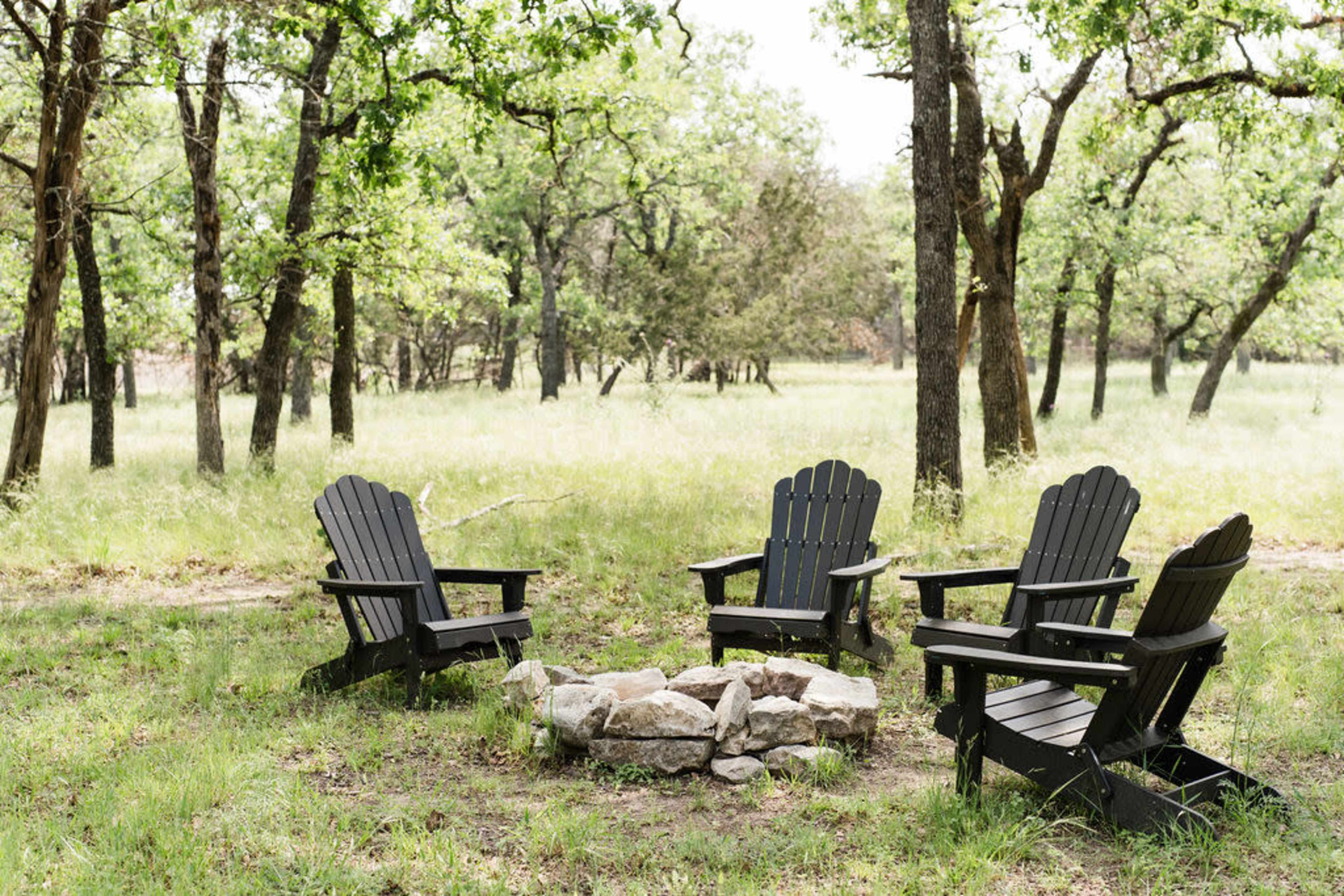 A circular arrangement of four black adirondack chairs surrounds a fire pit made of stones in a wooded clearing.