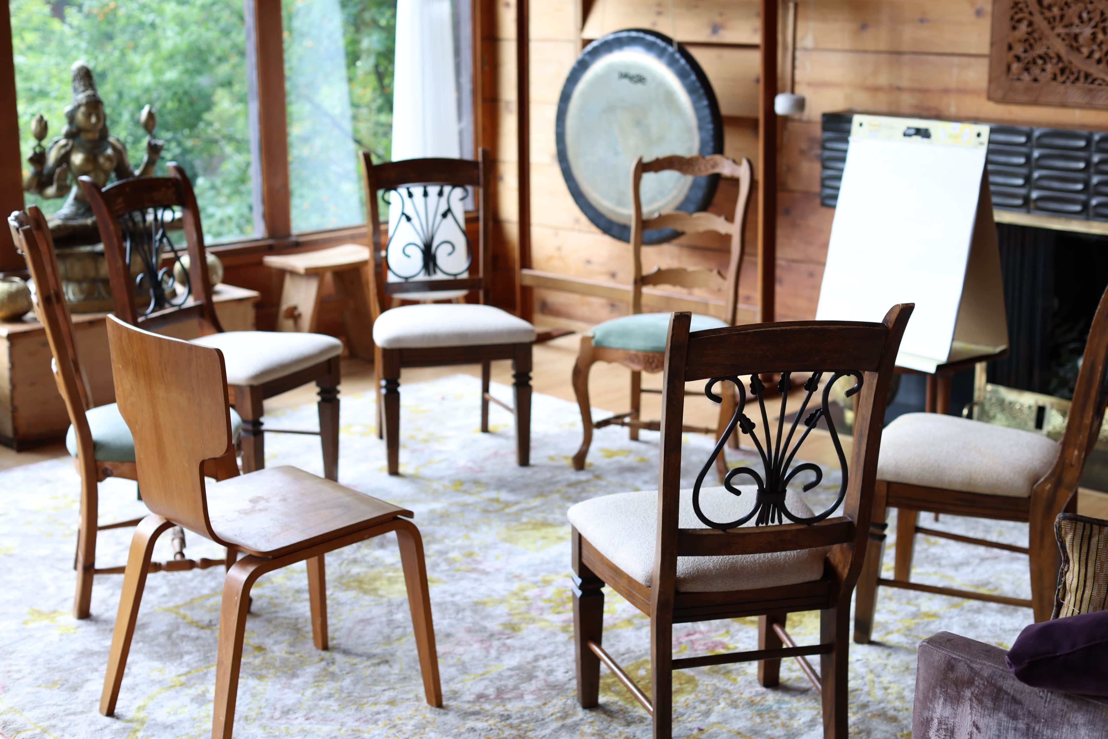 A circular arrangement of wooden chairs is set on a patterned rug in a room with wooden walls and large windows.