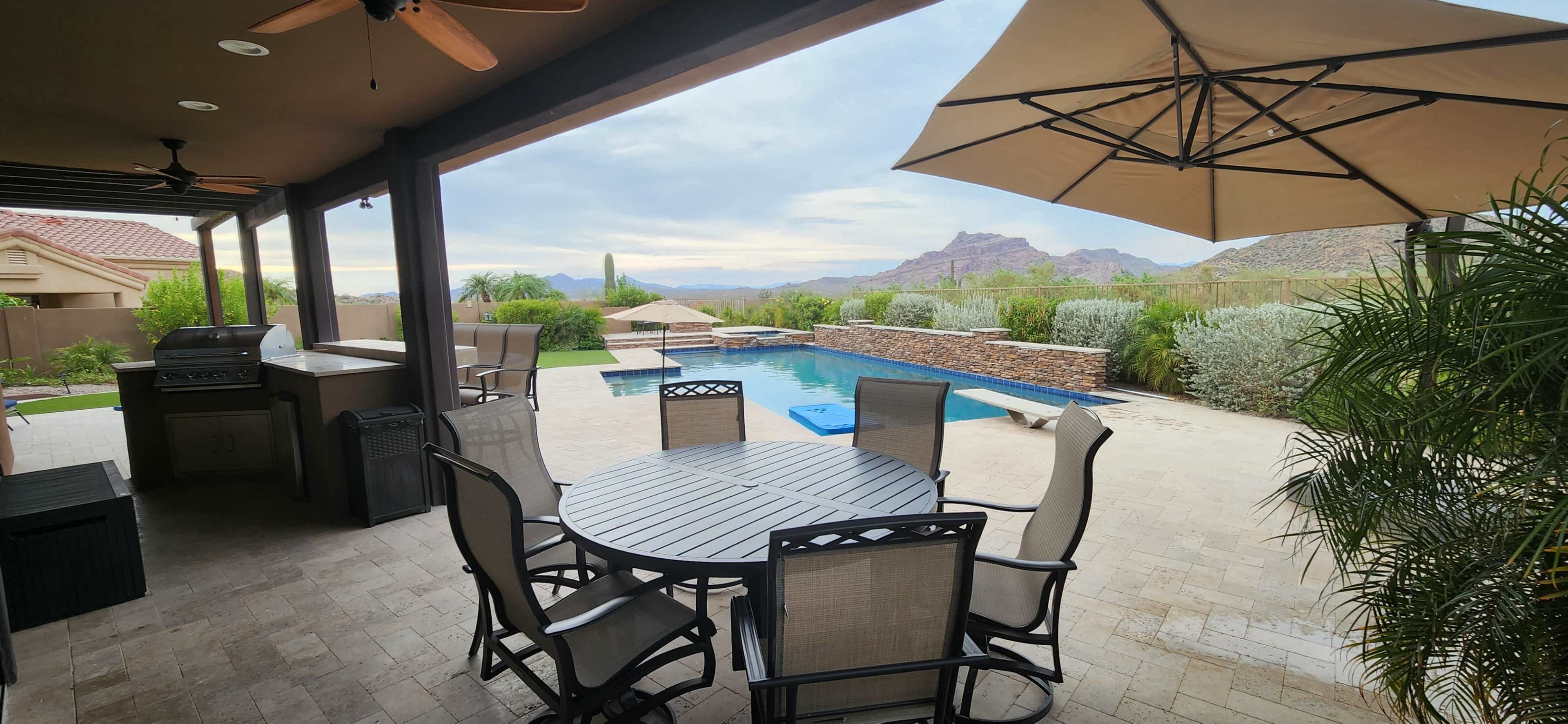 The image shows an outdoor patio with a round table and chairs, a swimming pool, and mountains in the background.