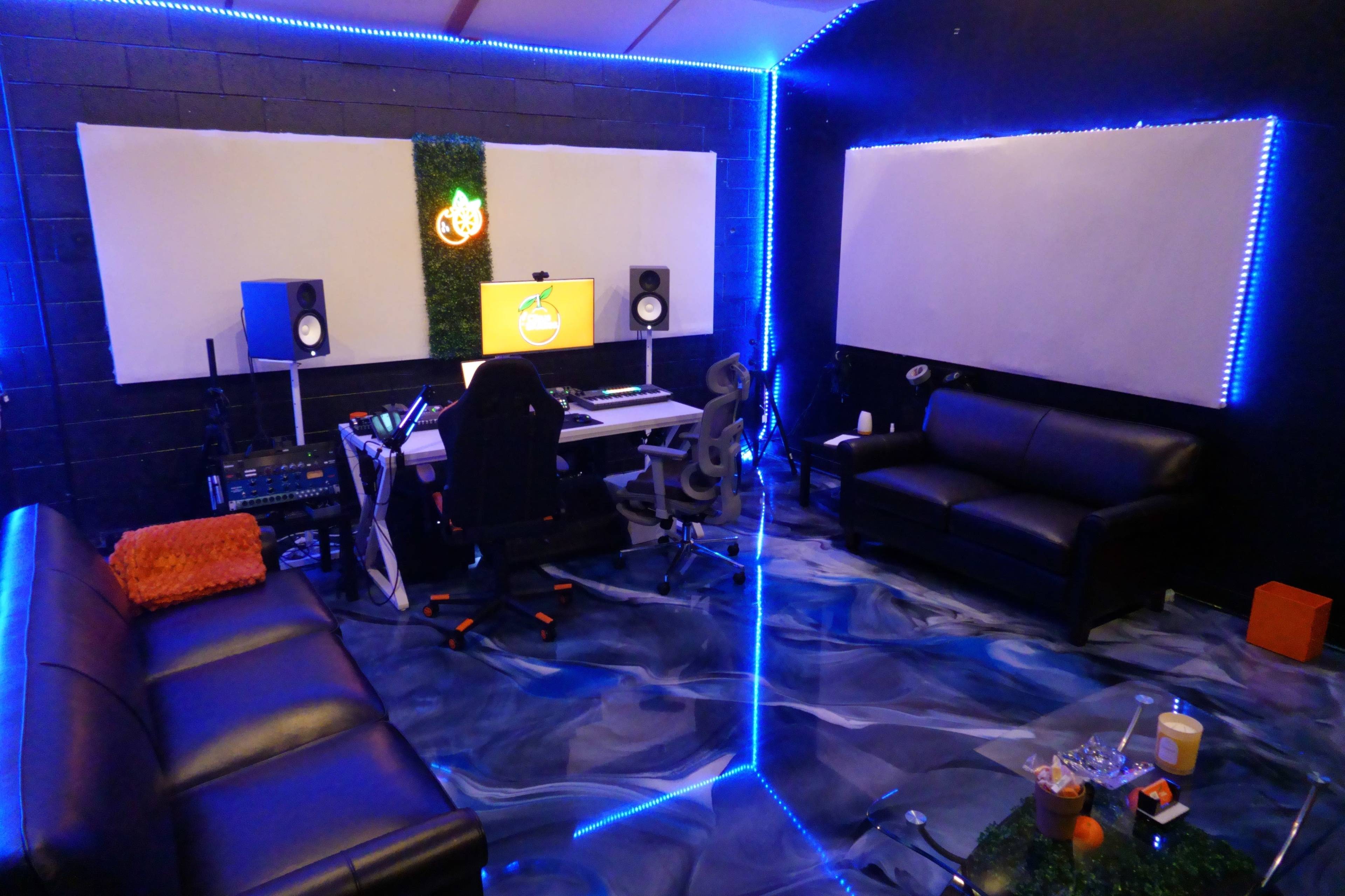 The image shows a modern music studio with a computer desk, speakers, a black couch, and blue LED lighting.