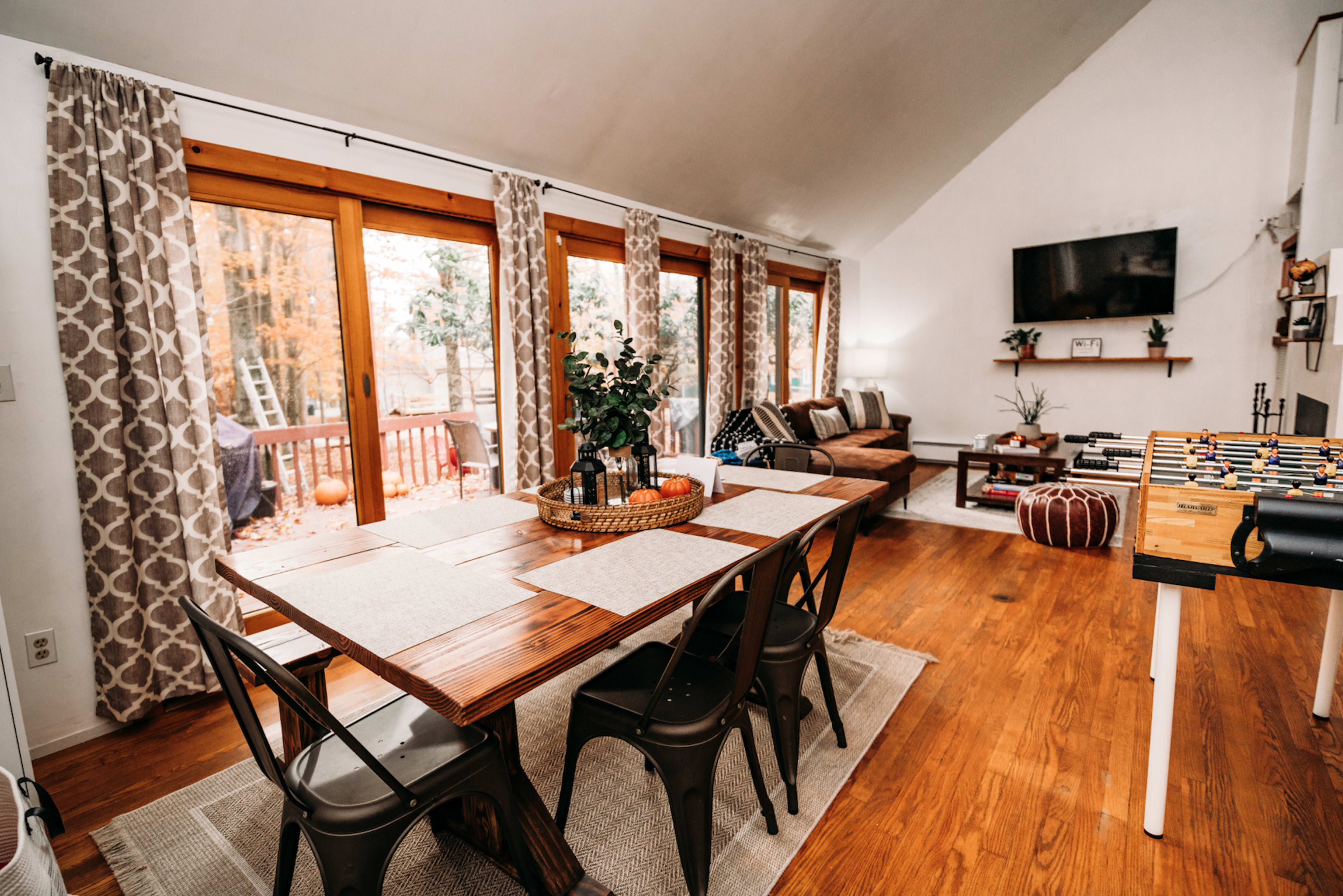 A dining area with a wooden table and black chairs, large windows allowing natural light, and a cozy living room space beyond with a sofa and a television.
