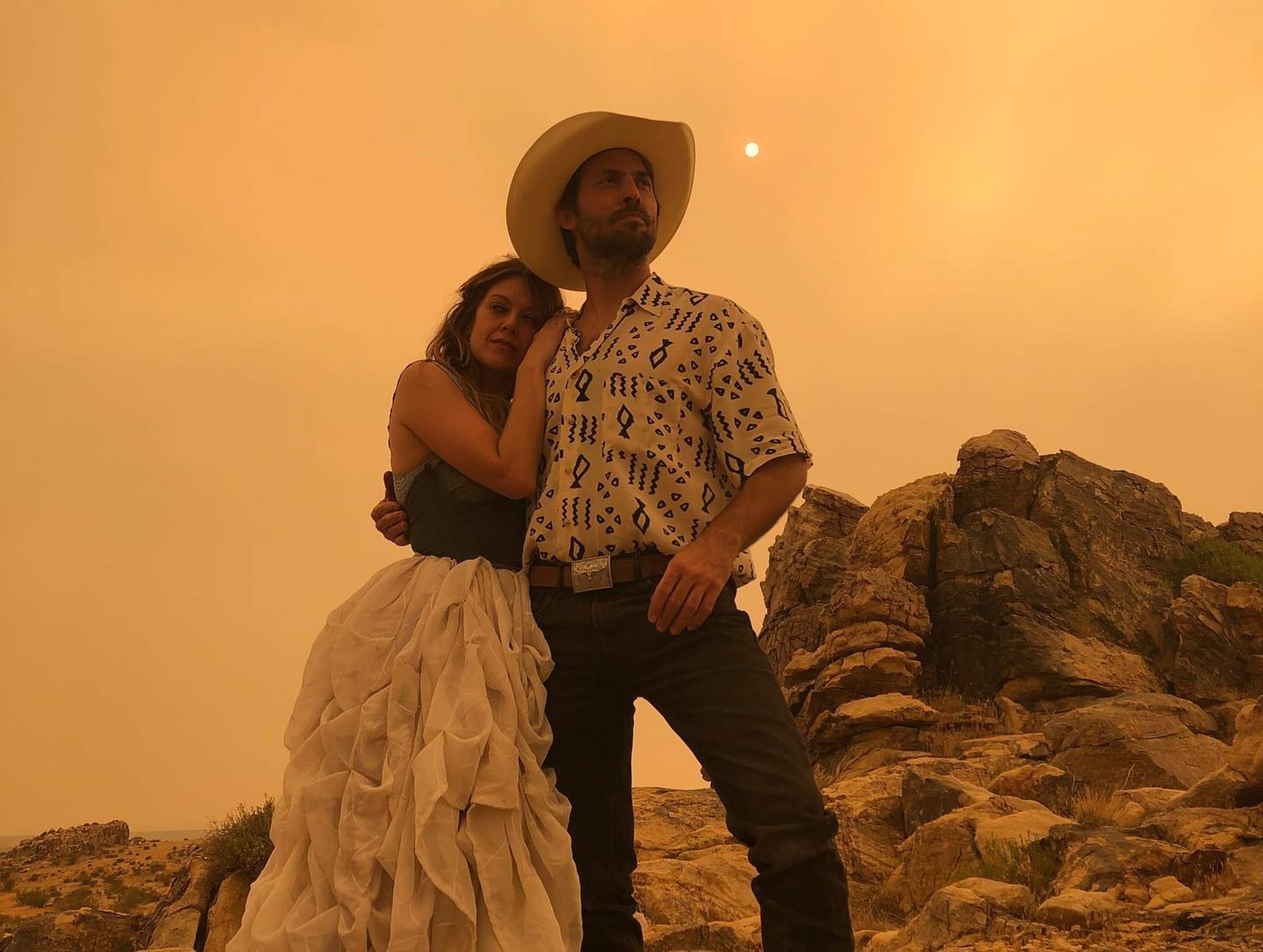A man in a cowboy hat and a woman in a flowing dress stand together on rocky terrain under an orange sky.