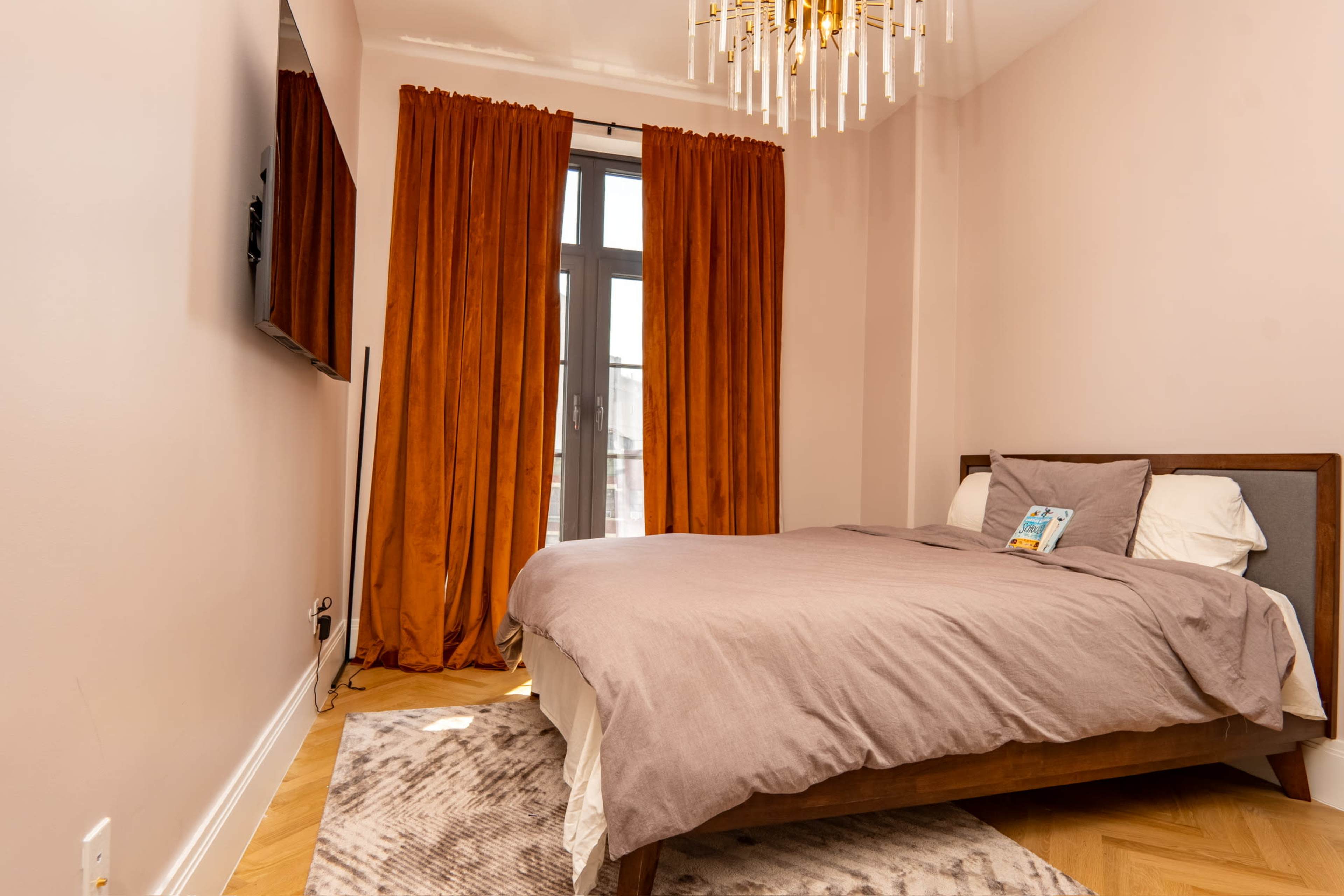 A bedroom features a bed with neutral bedding, a large window draped with orange curtains, a wall-mounted TV, and a patterned rug on the floor.