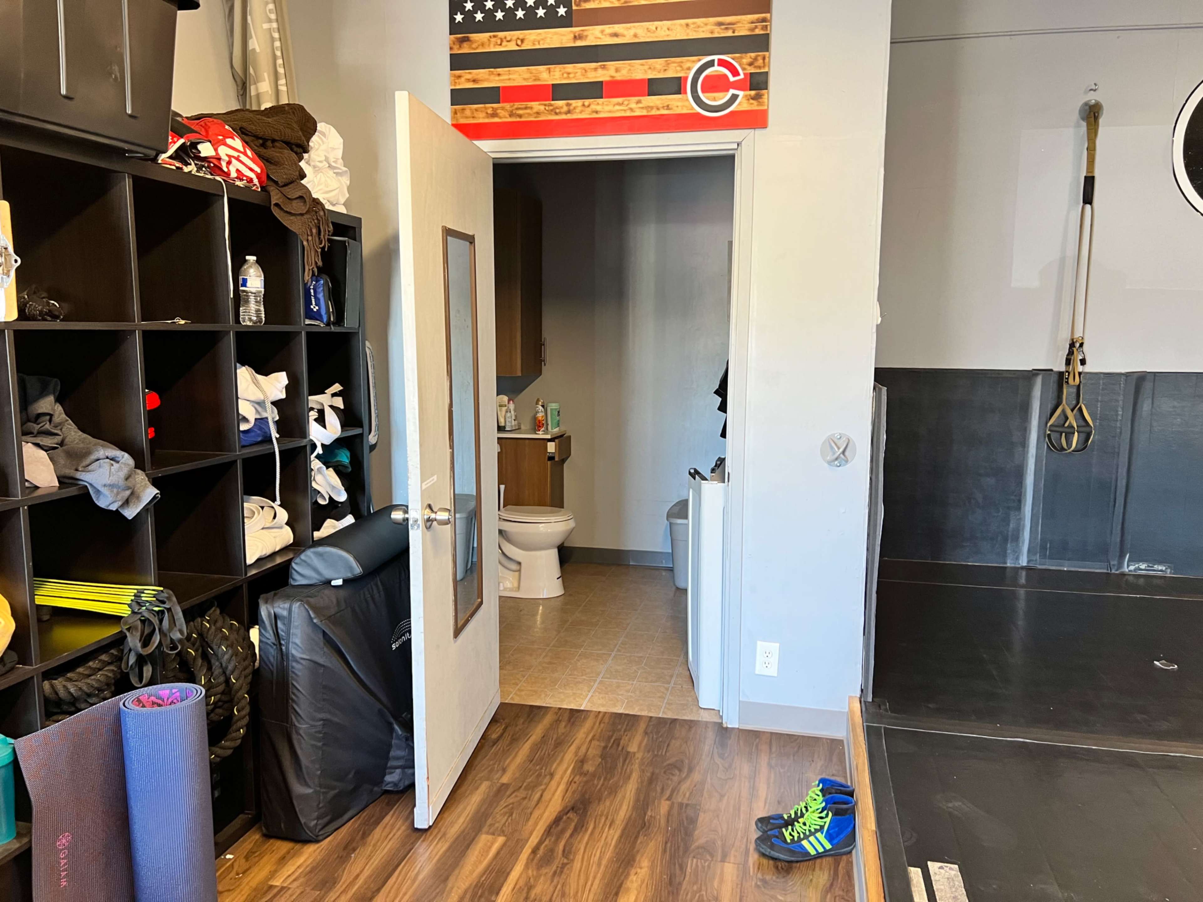 The image shows a room with a storage shelf filled with clothing, a partially open door leading to a restroom, and exercise equipment on the floor.