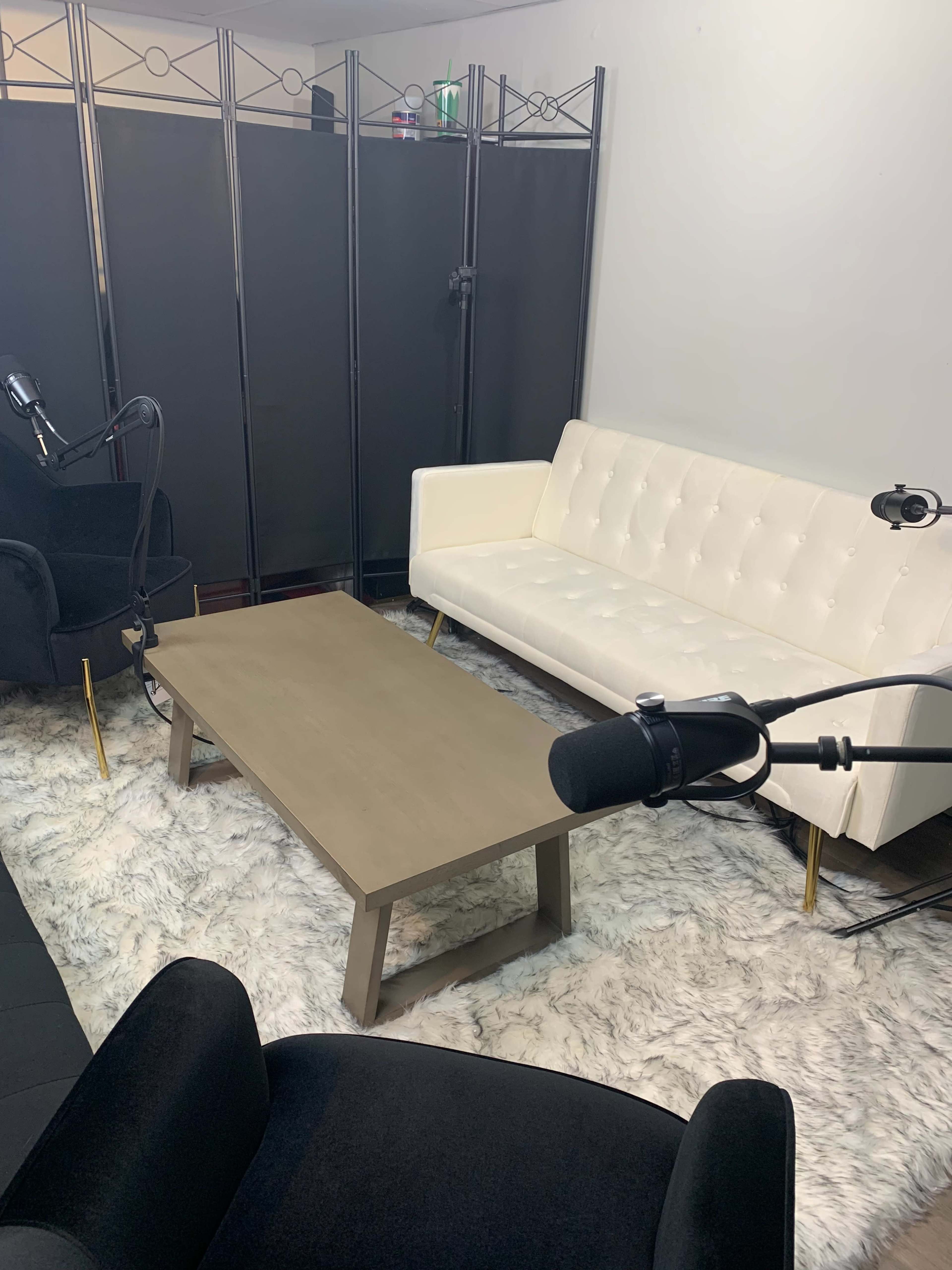 A minimalist recording studio setup with a white sofa, a wooden coffee table, and two microphones positioned near black chairs on a fluffy rug.