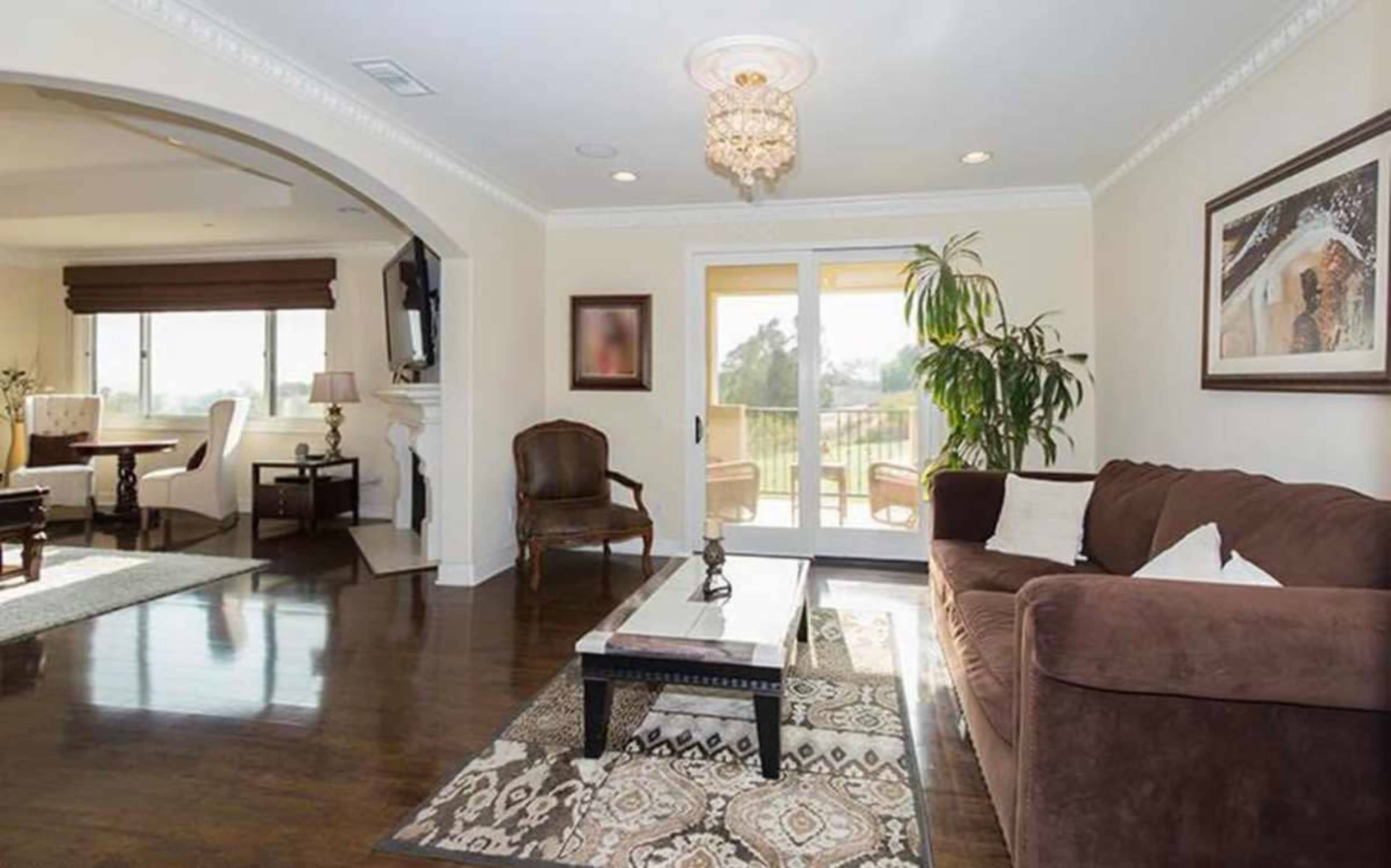 The image shows a well-lit living room with a brown sofa, a coffee table on a patterned rug, and large windows that open to a balcony, while a dining area is visible in the background.