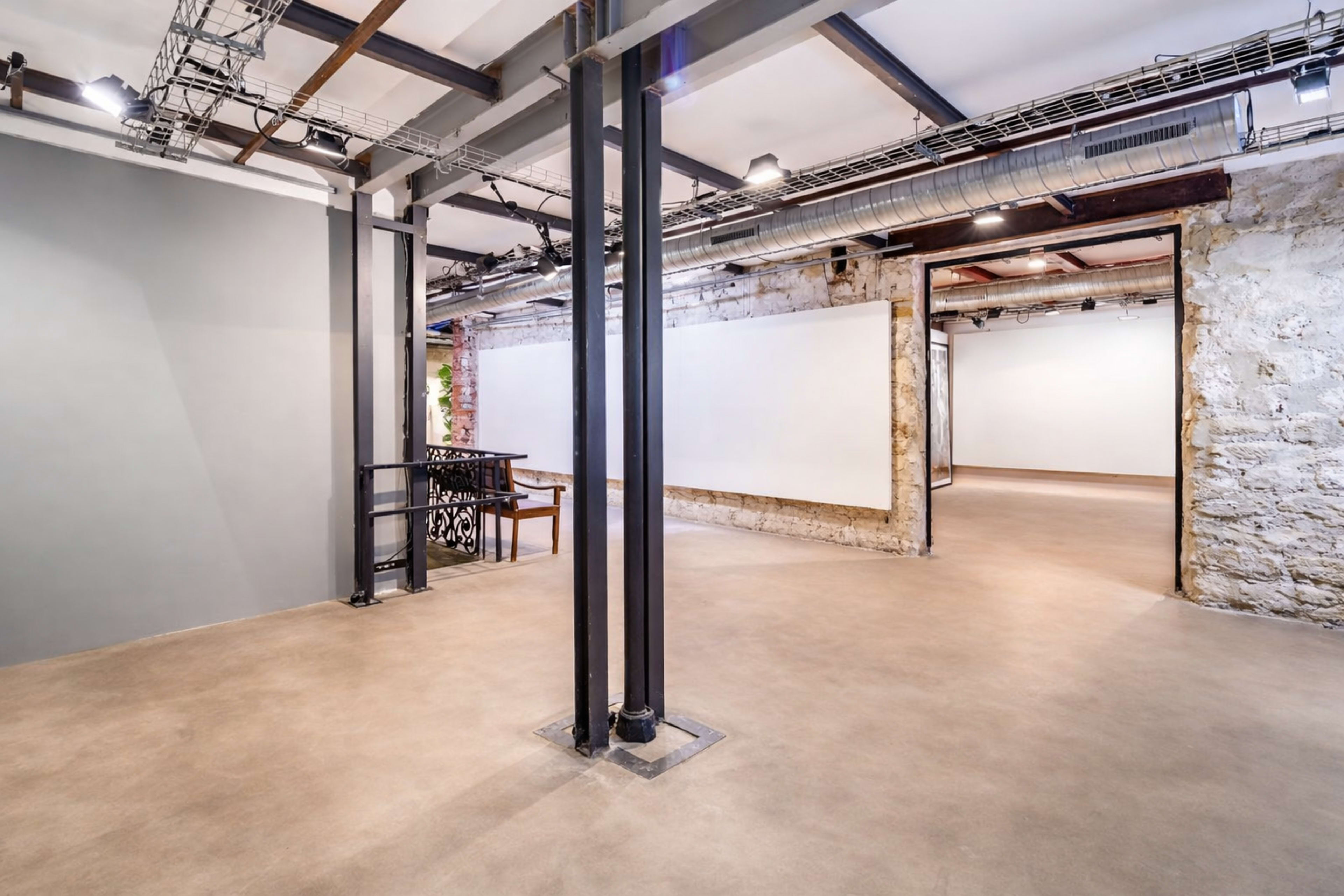 The image shows an interior space featuring a large, open area with exposed brick walls, metal pillars, and white walls suitable for displaying artwork.