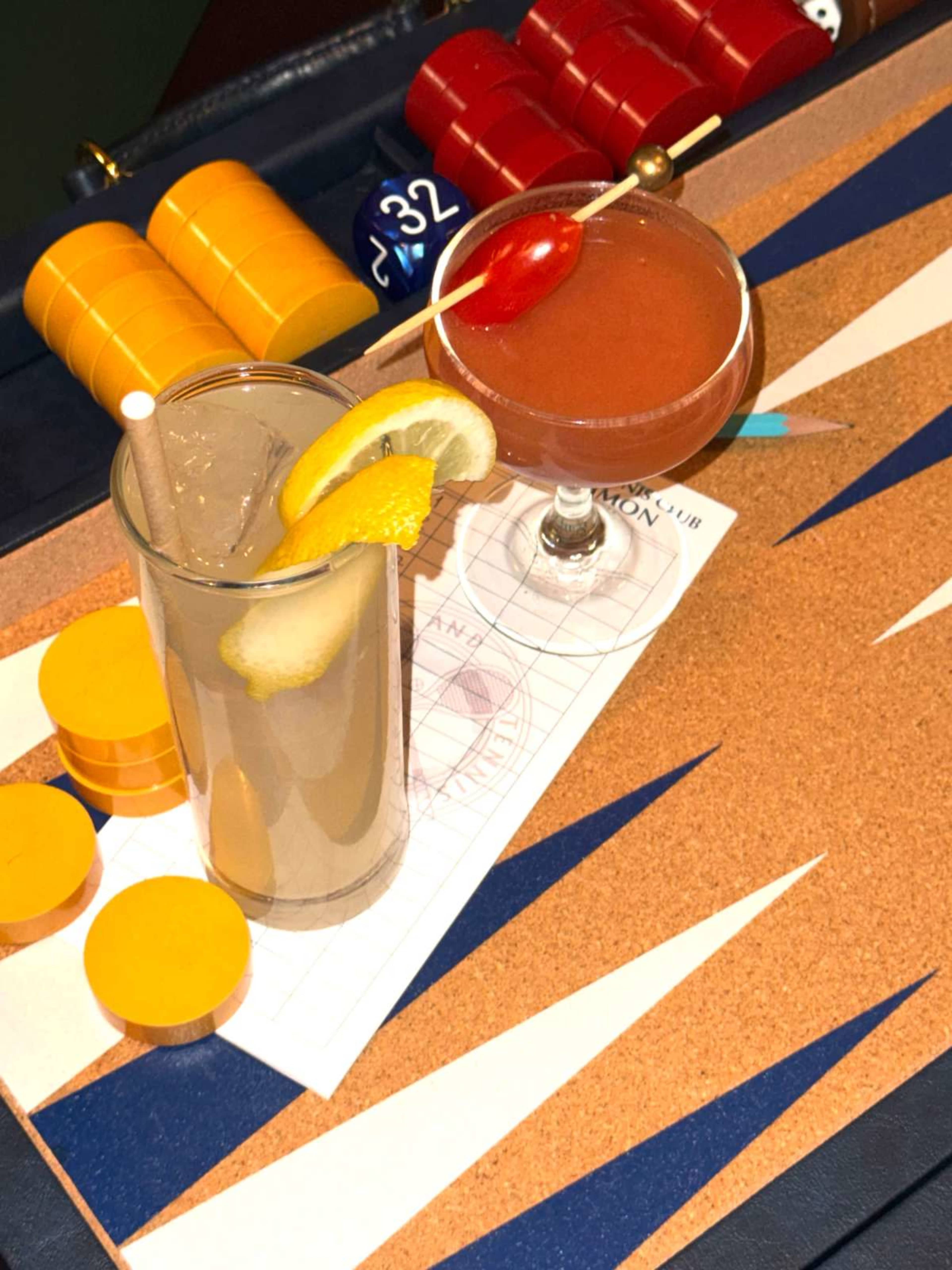Two cocktails are placed on a cork background, with a tall glass containing a lemon garnish and a short glass decorated with a cherry, alongside colorful game chips.