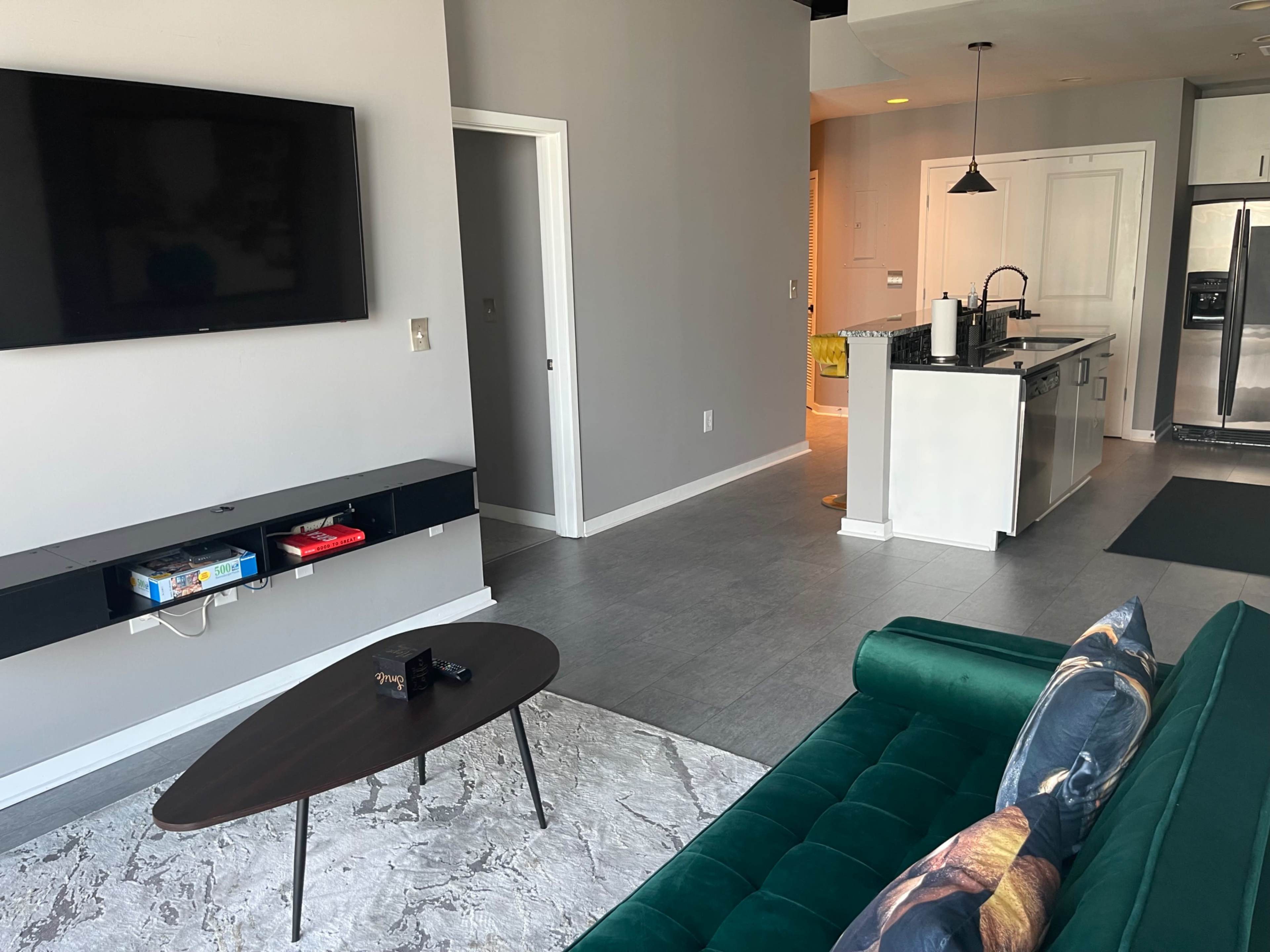 The image shows a modern living room with a green sofa, a coffee table, a wall-mounted TV, and an open kitchen in the background.